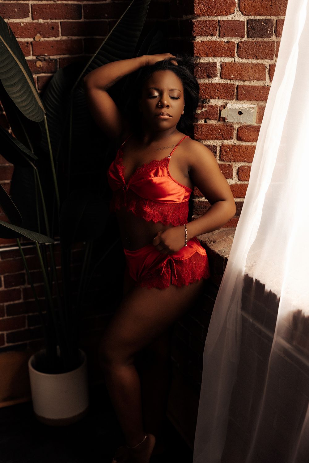 Woman in red lingerie posing gracefully against a brick wall with sheer curtains and tropical plant.