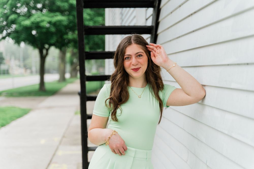 A person in a mint green outfit poses casually against white siding near outdoor stairs.
