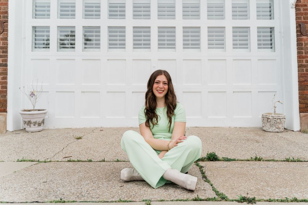 A person in a light green outfit sits cross-legged on concrete in front of a white garage door.