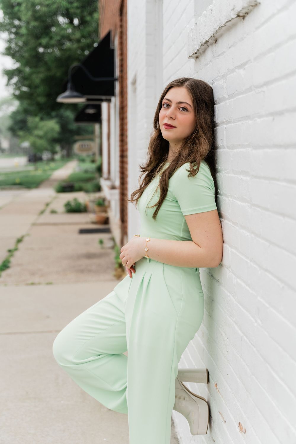 A person in mint green attire poses against a white brick wall with trees in background.
