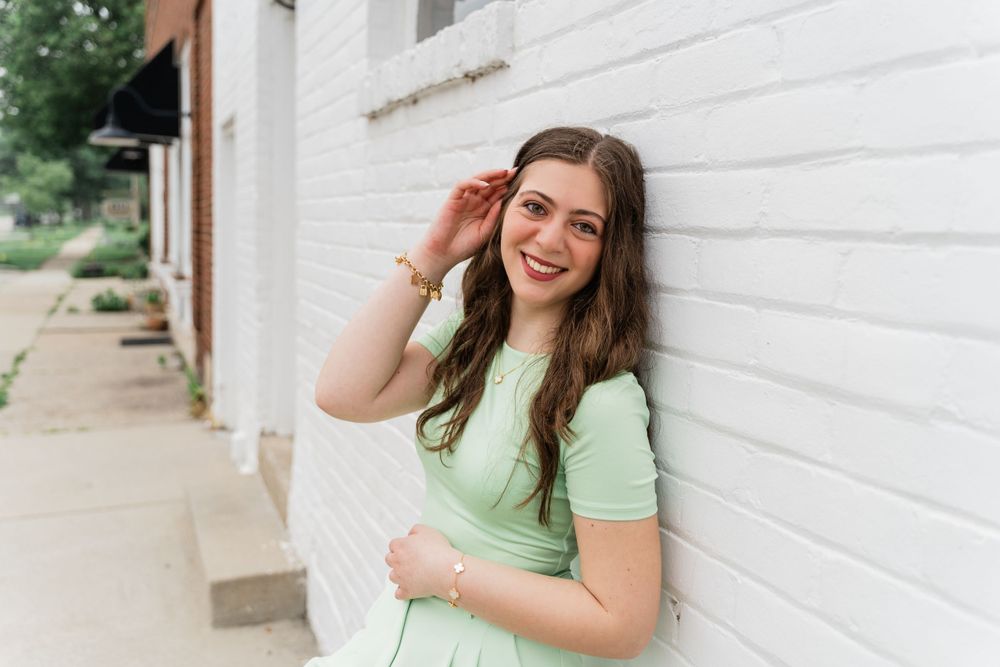 A person in mint green attire touching their hair while posing against a white wall.