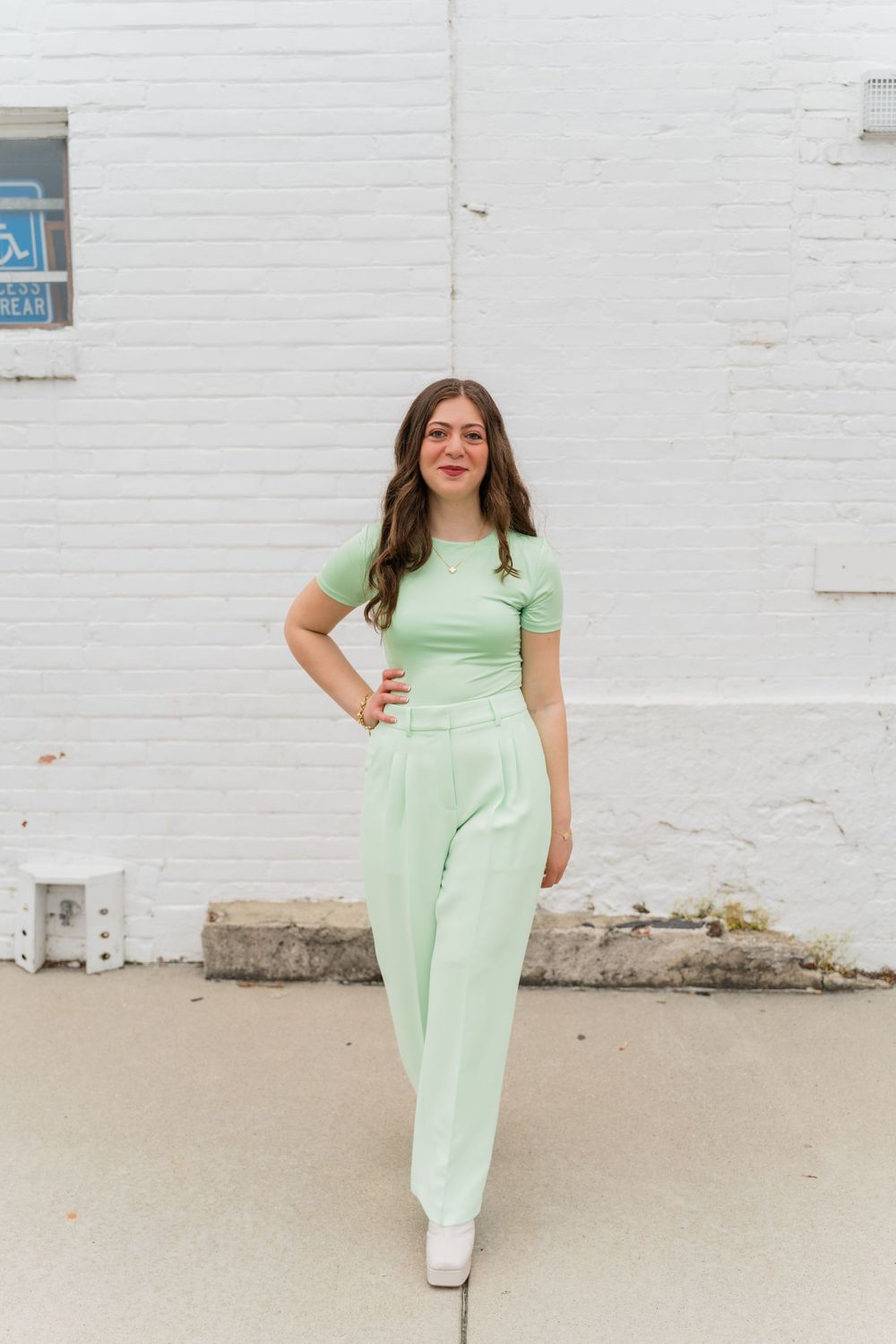 A full-length portrait of someone in a pastel green jumpsuit against a white brick wall.