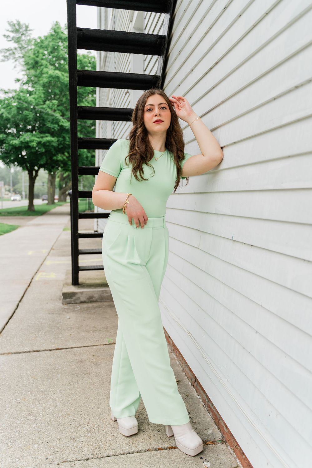 A person in mint green poses near black stairs against white siding.