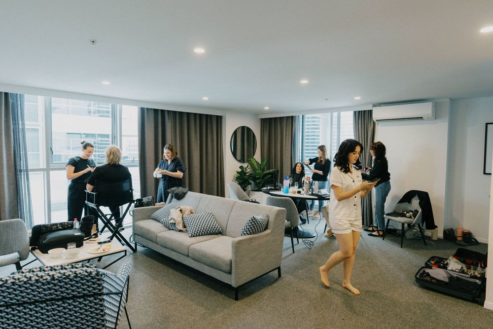 People getting ready in a modern hotel room with gray couch and floor-length curtains.