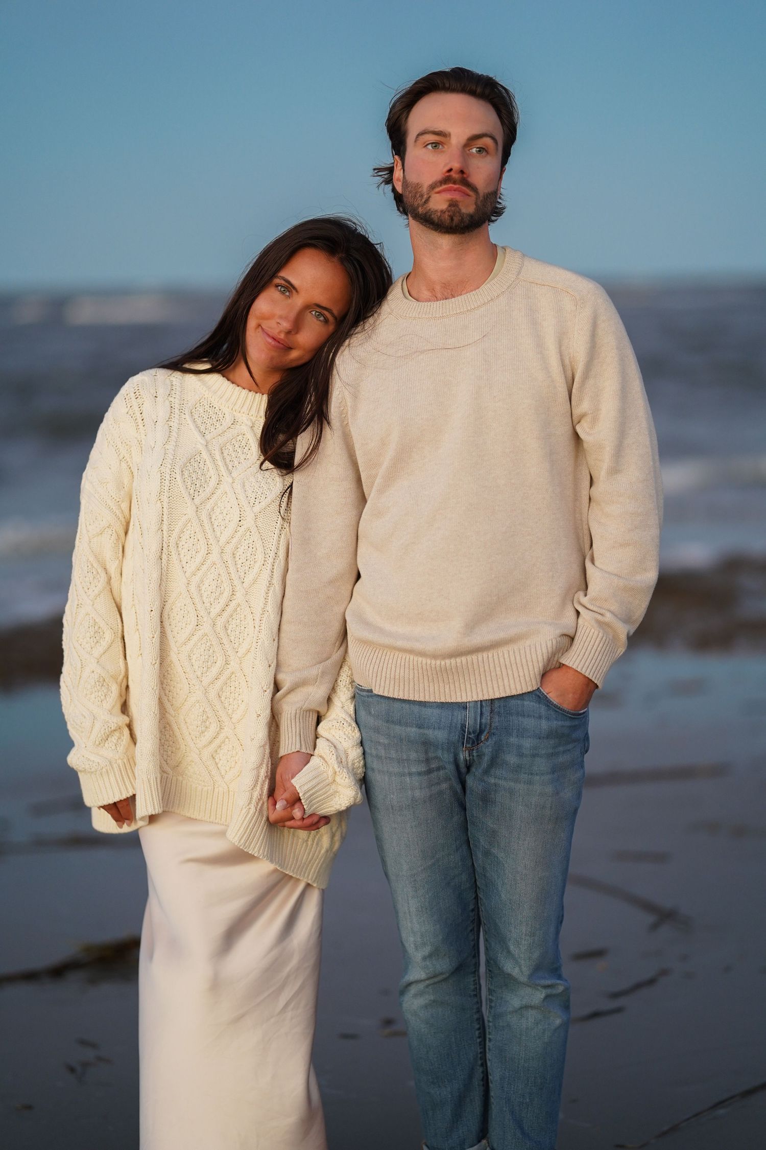 Couple in casual sweaters and jeans pose together on a beach at sunset.