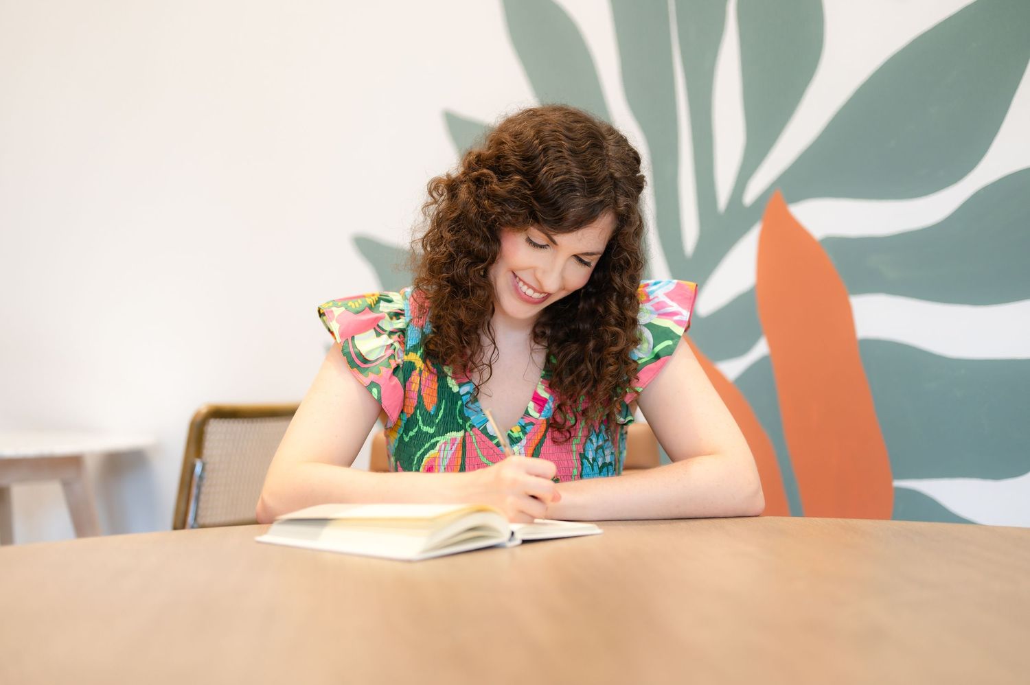 Person with curly hair reading book at wooden table with colorful wall art background.