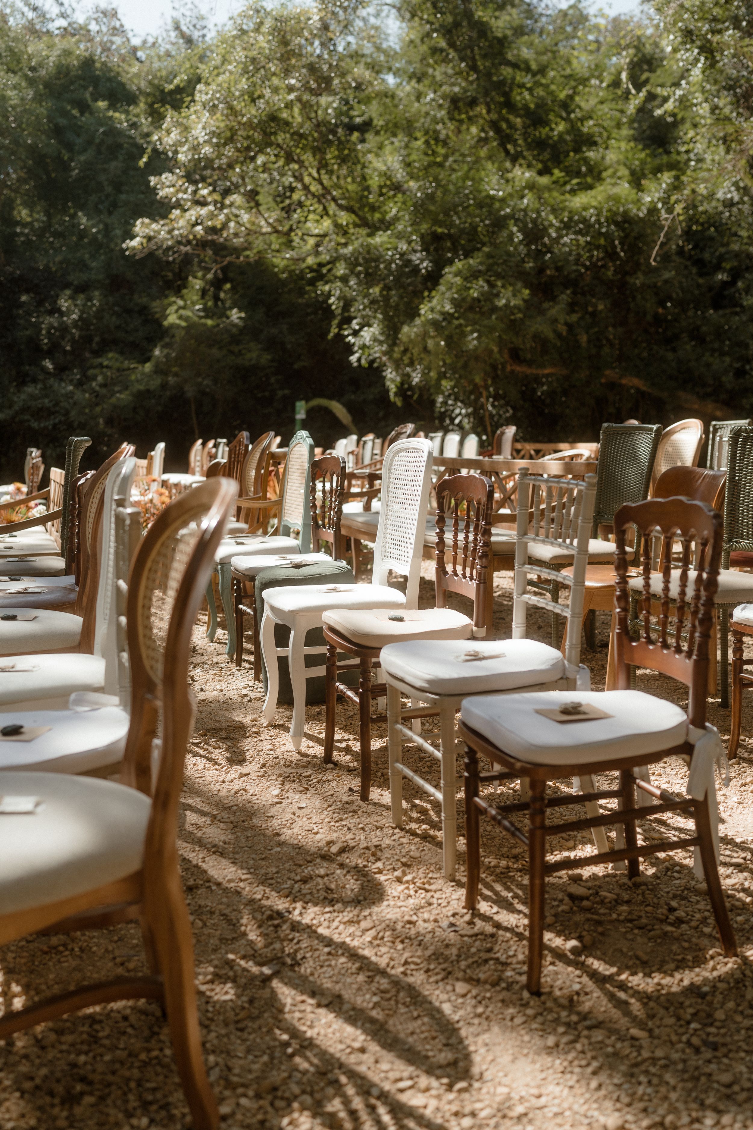 Rows of vintage wooden chairs arranged outdoors under shady trees for a wedding ceremony.