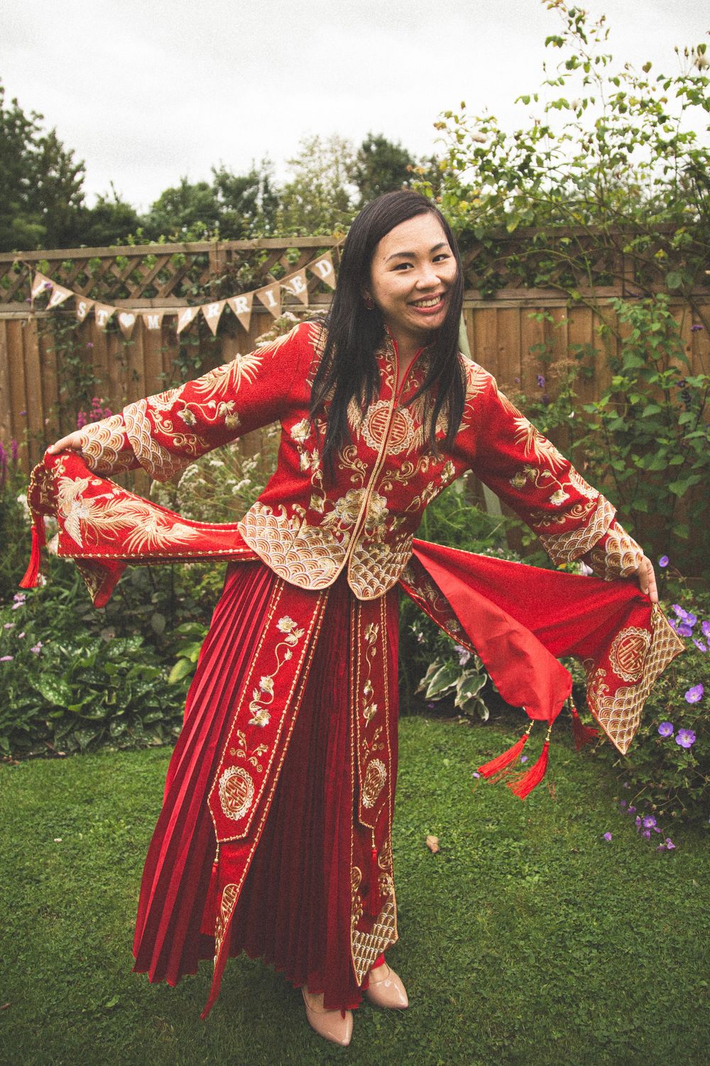 Bride twirls in ornate red and gold traditional Chinese wedding dress in garden setting.