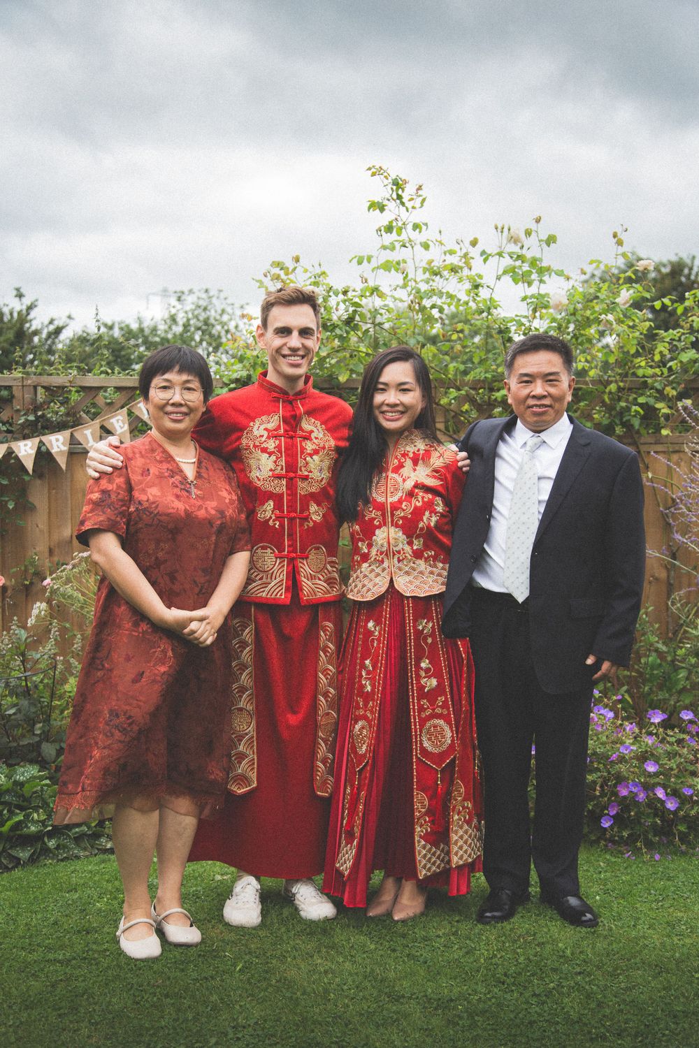 Wedding group photo featuring blend of Western and traditional Chinese red wedding attire in garden.