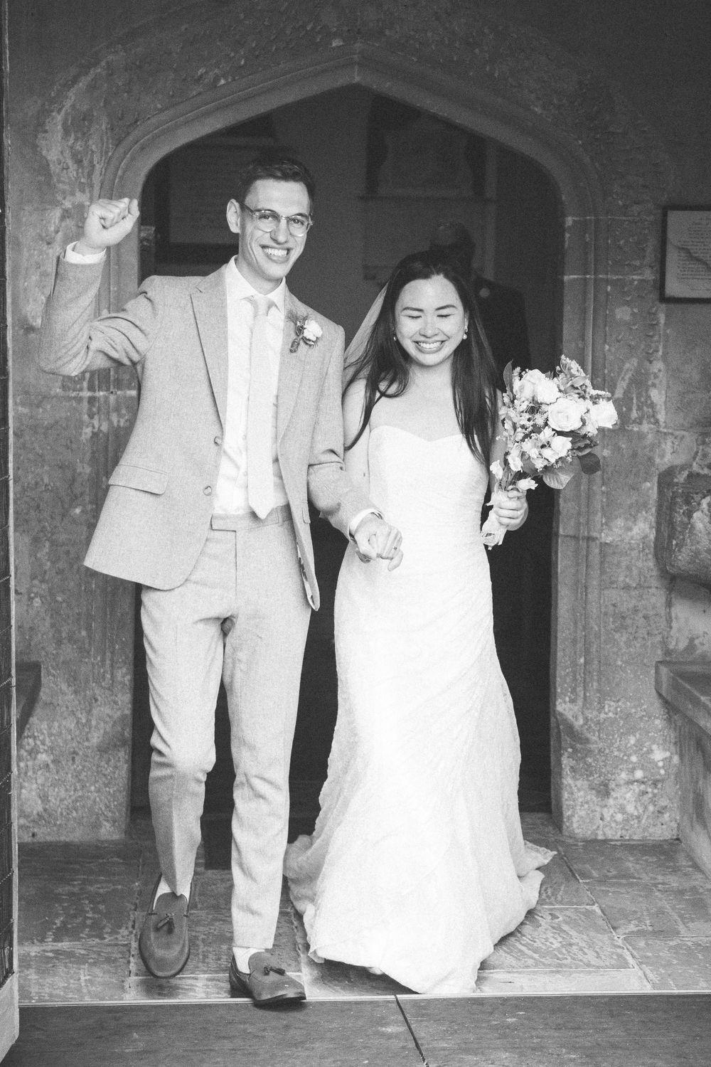 Newlyweds celebrate with joy as they exit a historic stone archway after their wedding ceremony.