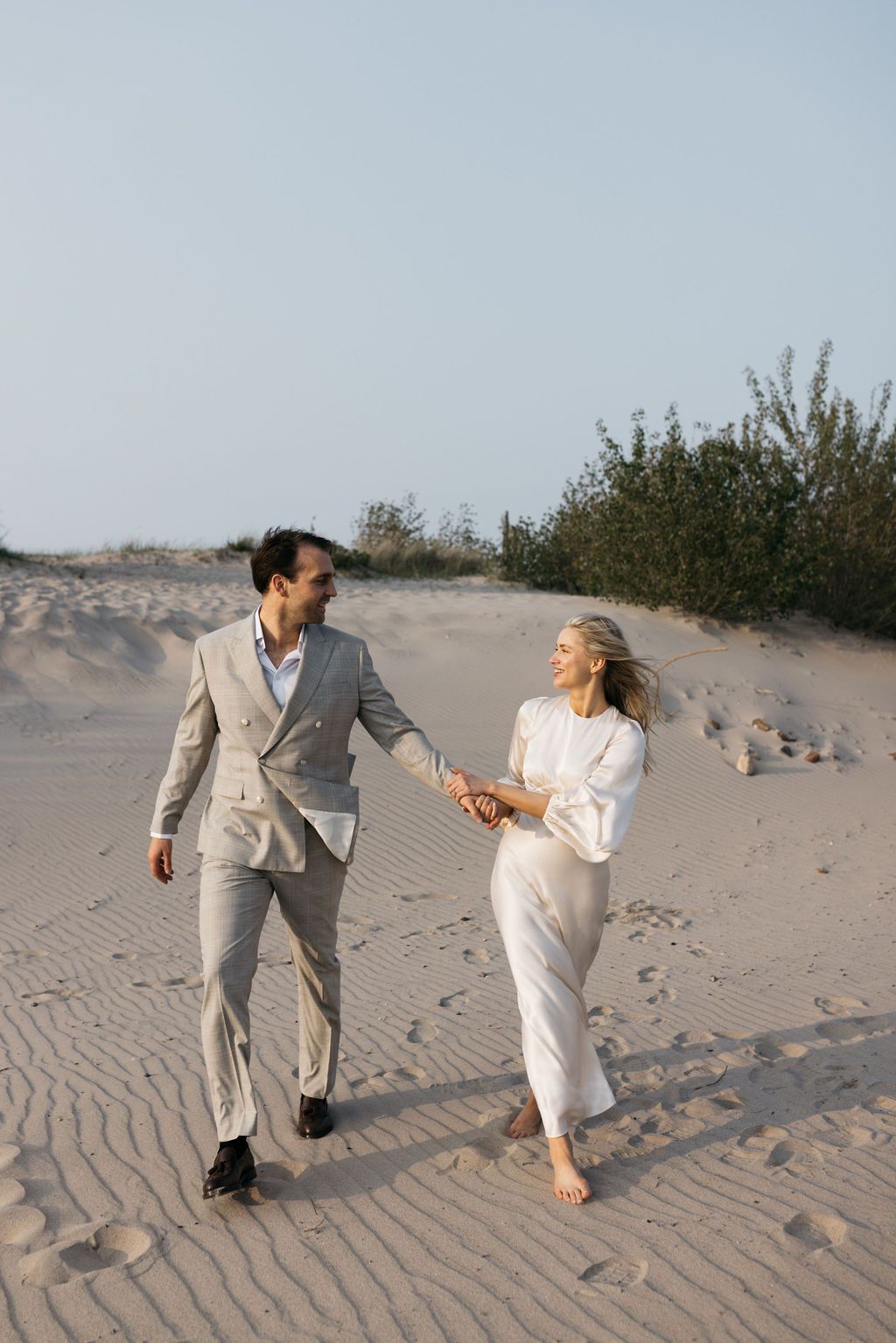 Couple walking hand in hand on sandy beach wearing light colored outfits.