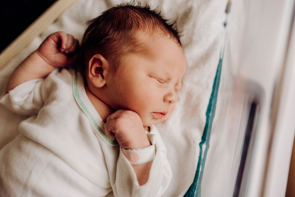 Sleeping newborn in white onesie lying on soft white bedding.