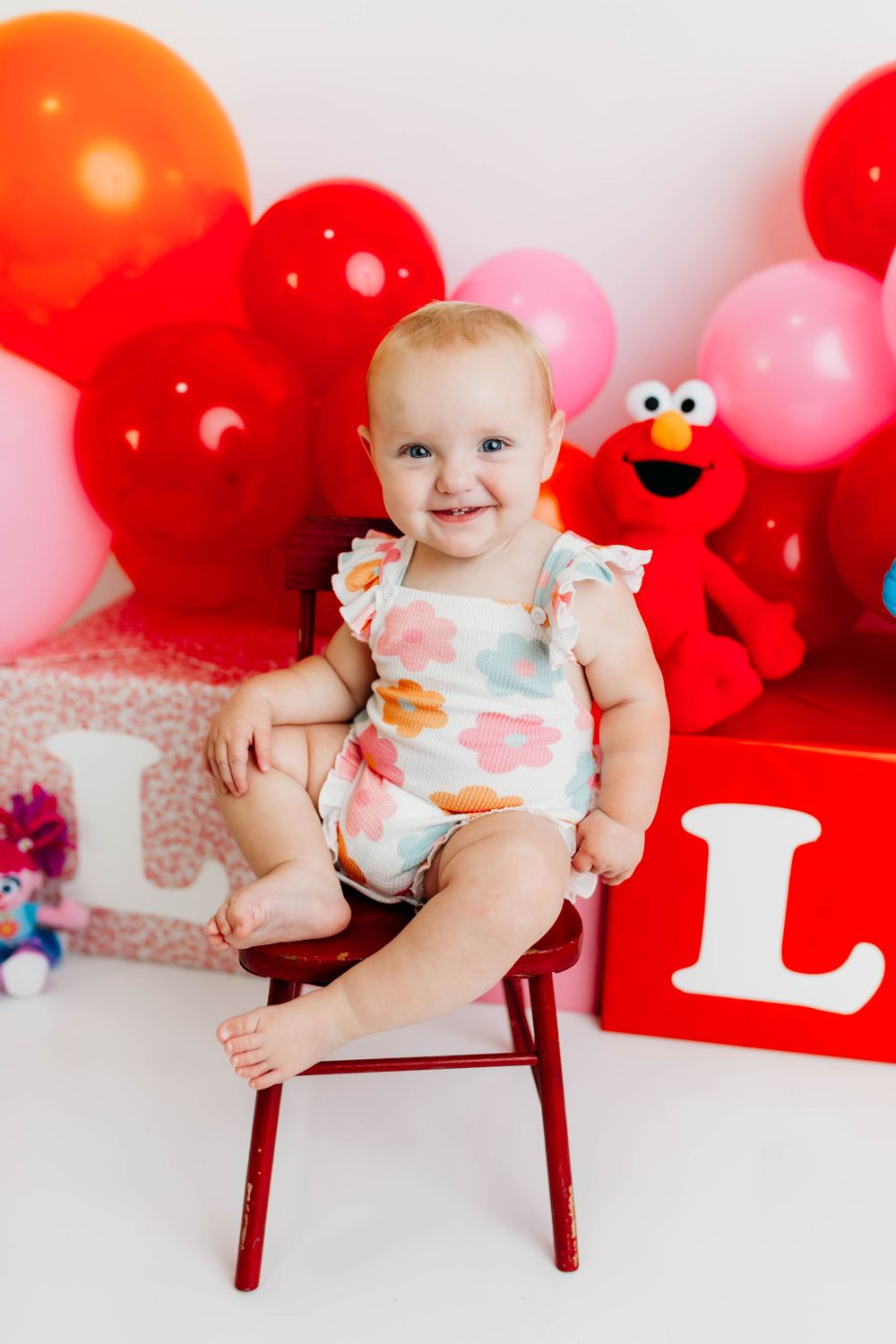 Baby celebration setup with red and pink balloons, Elmo plush toy, and decorative letter L.
