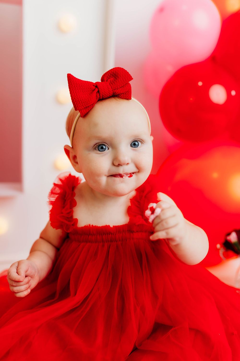 Baby wearing red dress with bow and sparkly holiday decorations in background.