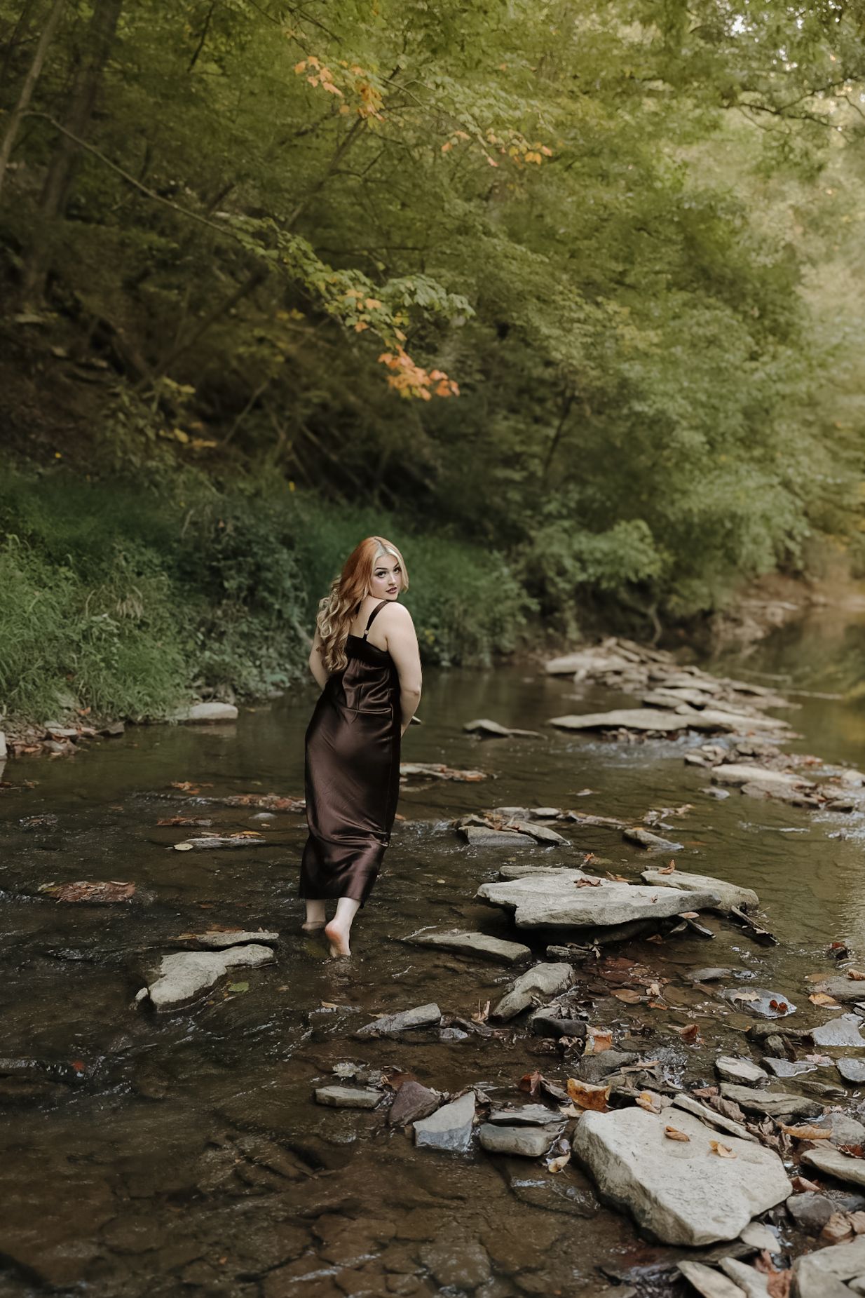 Woman in black dress stands barefoot in rocky stream surrounded by lush forest.