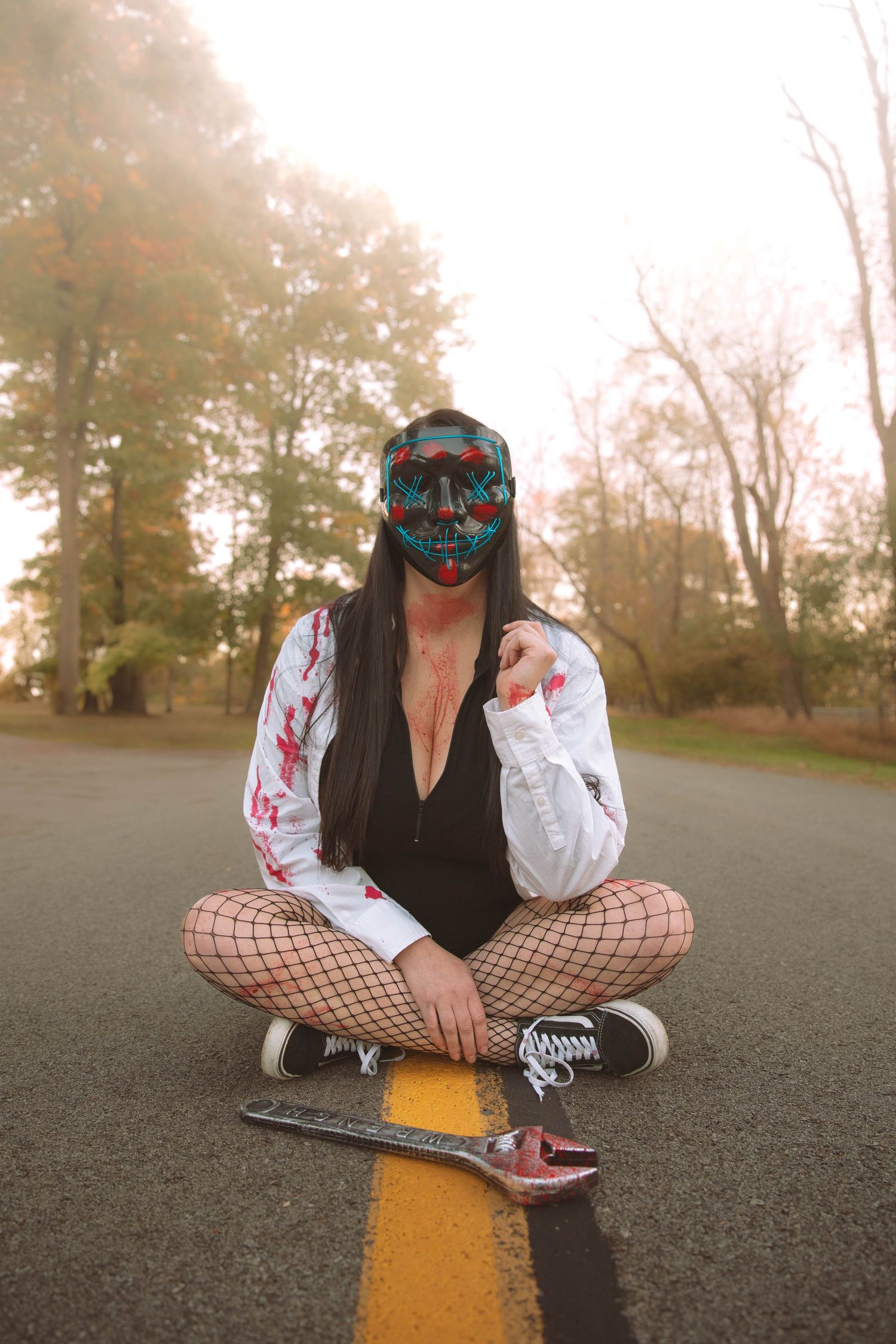 Person in Halloween costume with LED mask sitting on yellow road line with skateboard.