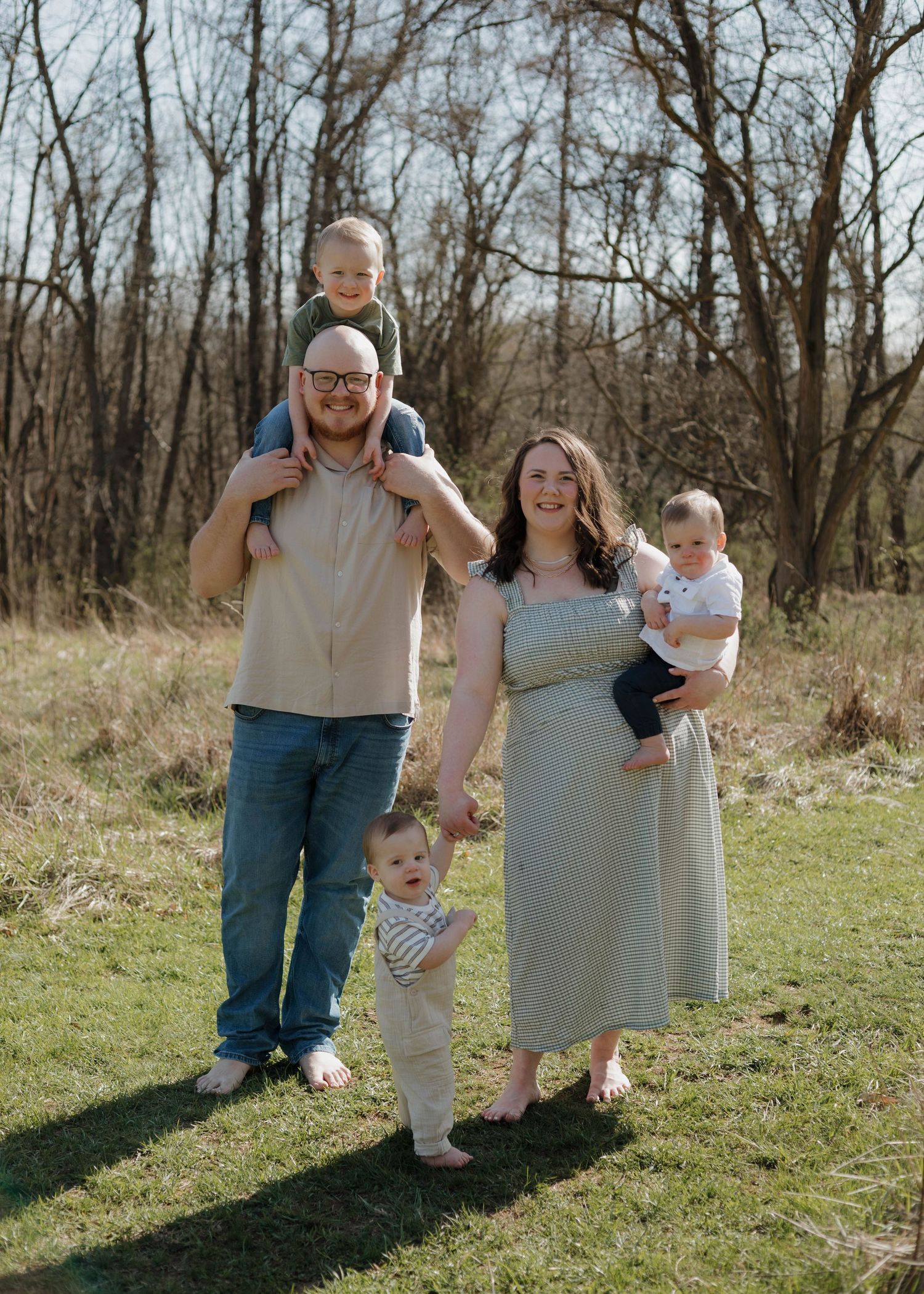 A family stands together in a grassy field on a sunny day with bare trees in the background.