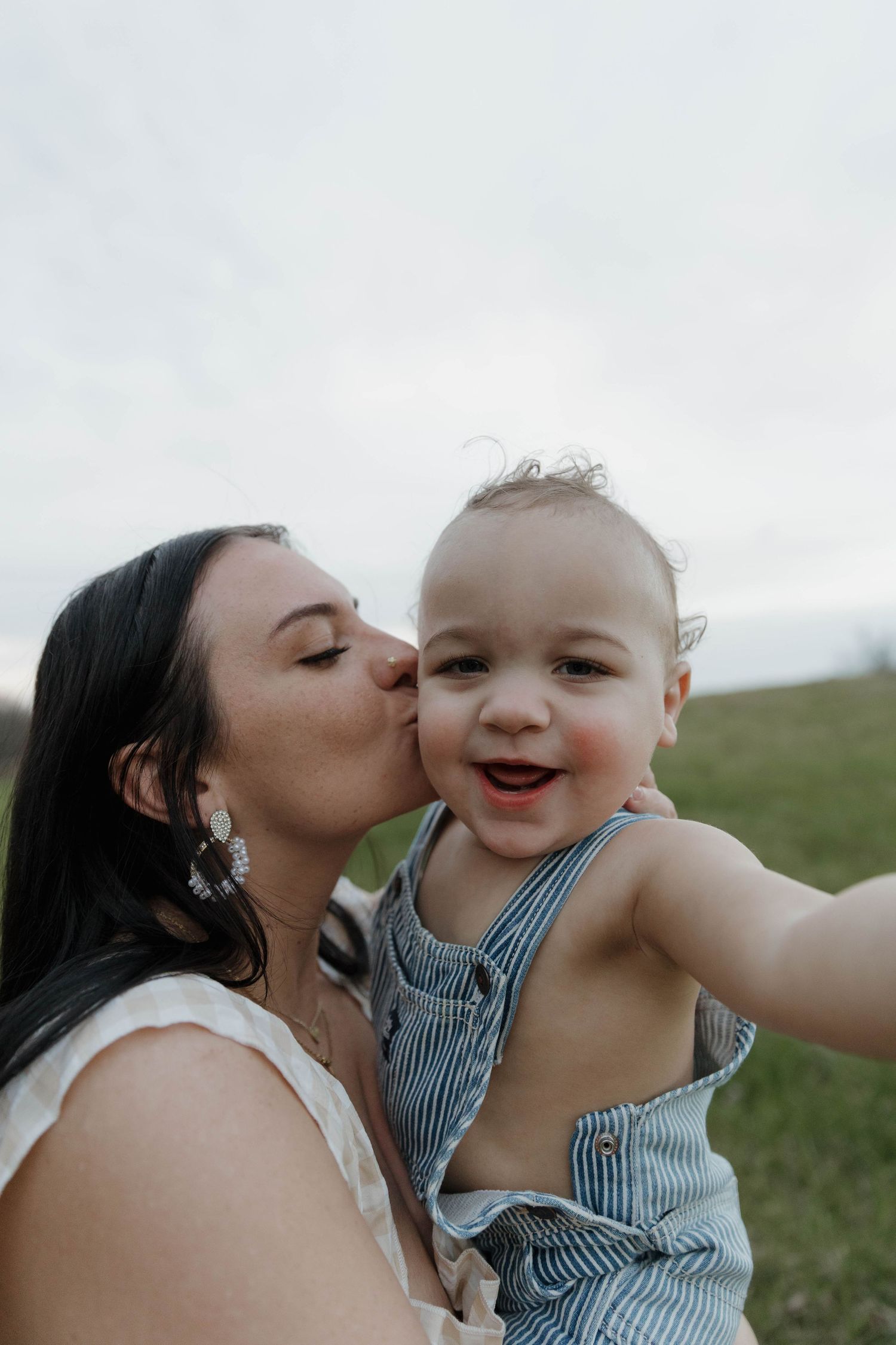 A tender moment outdoors as a parent kisses their happy toddler wearing blue and white striped overalls in a grassy field.