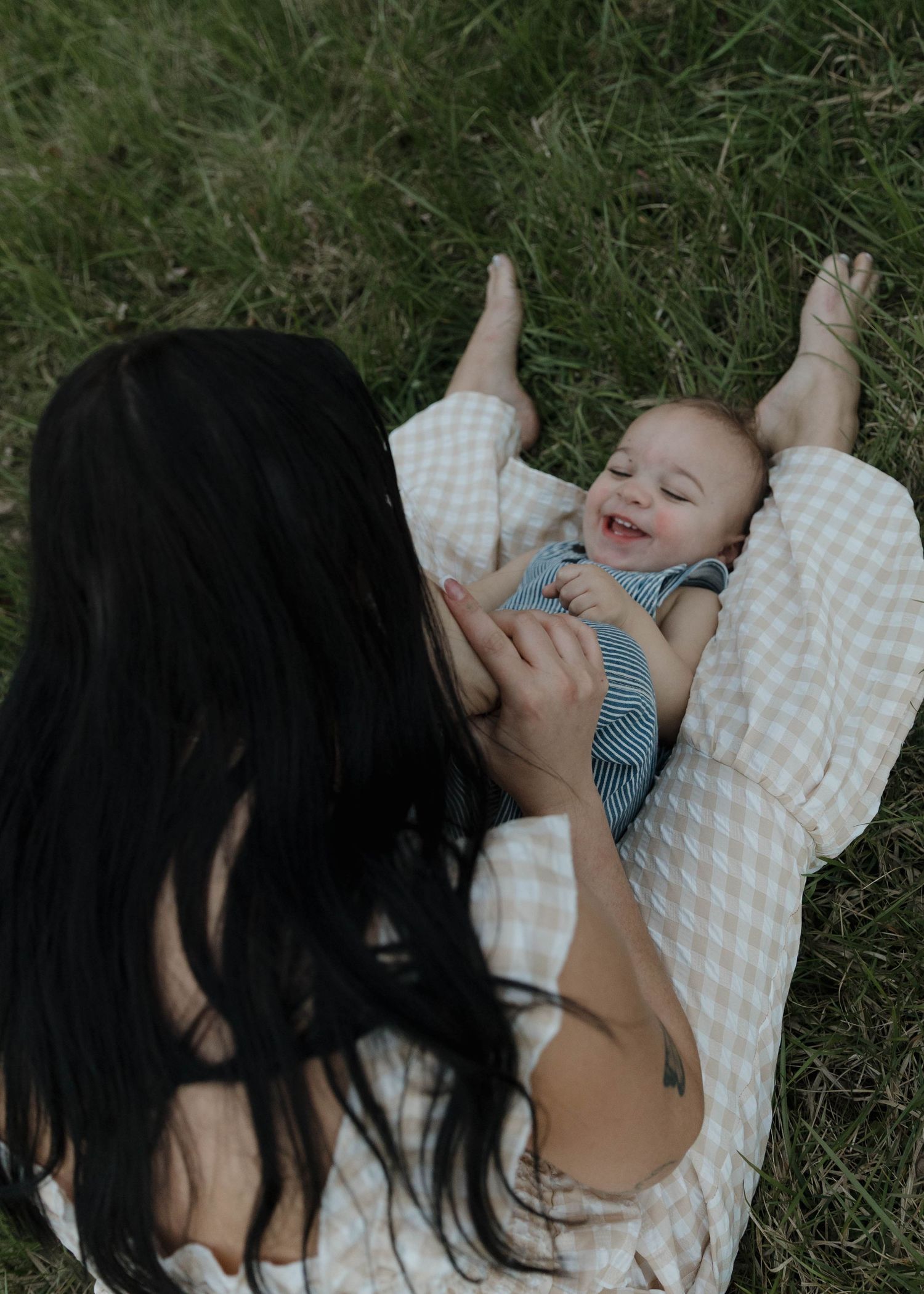 A mother in a checkered shirt plays with her laughing baby while laying together in green grass.