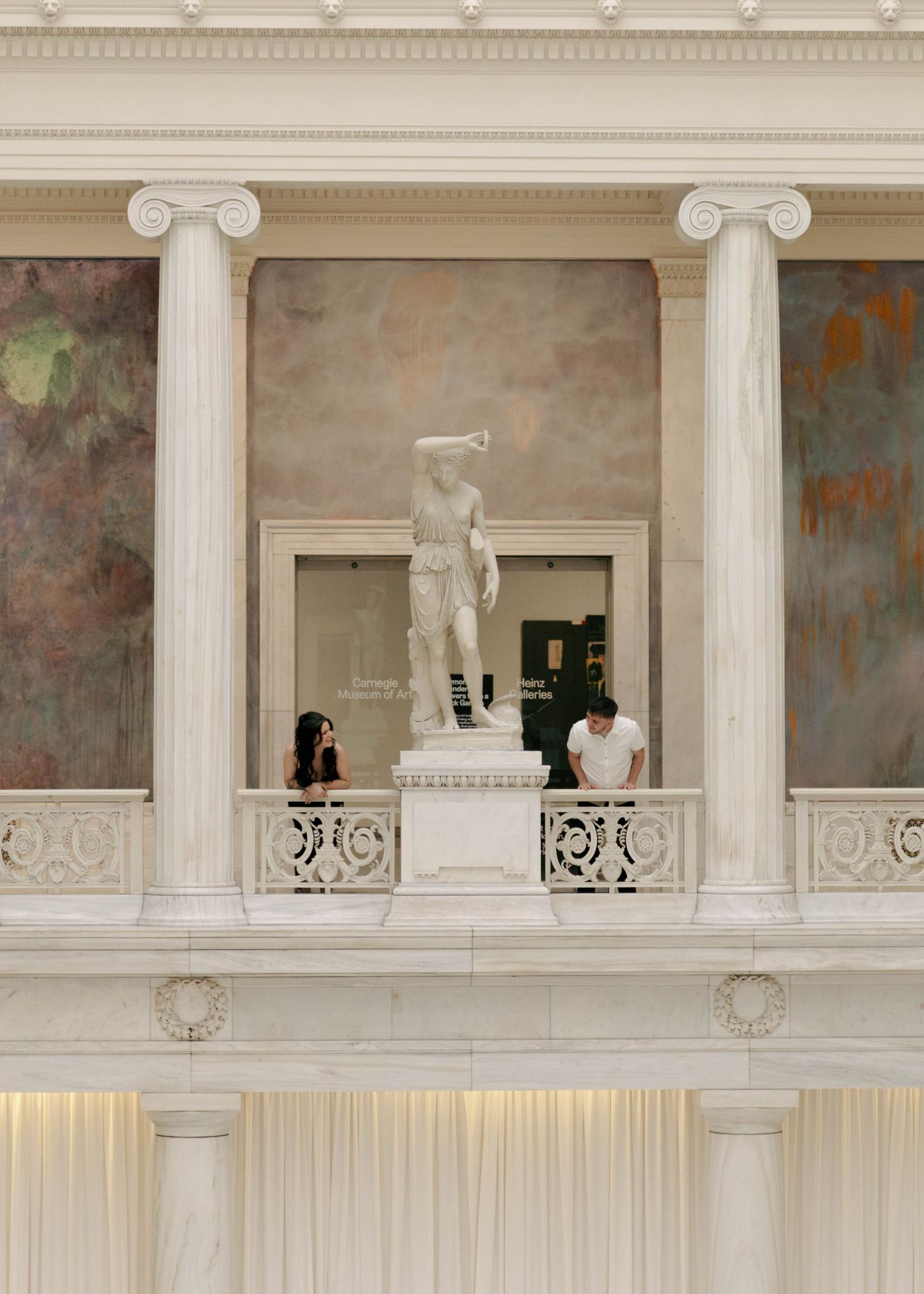 Classical museum interior with white Ionic columns framing a viewing balcony featuring a statue and draped curtains below.