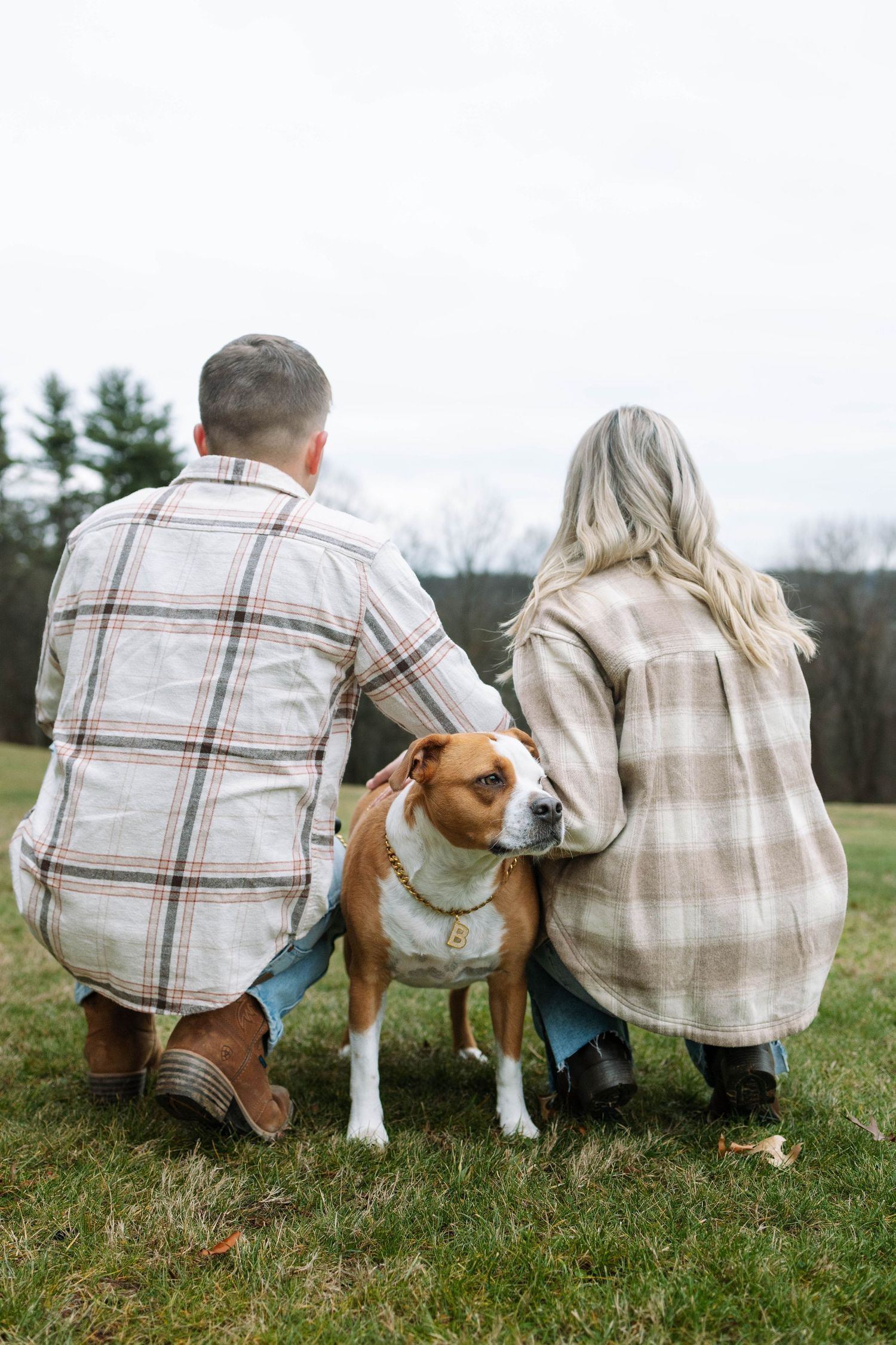 A couple in plaid shirts sitting on grass with their brown and white dog on a cloudy day.