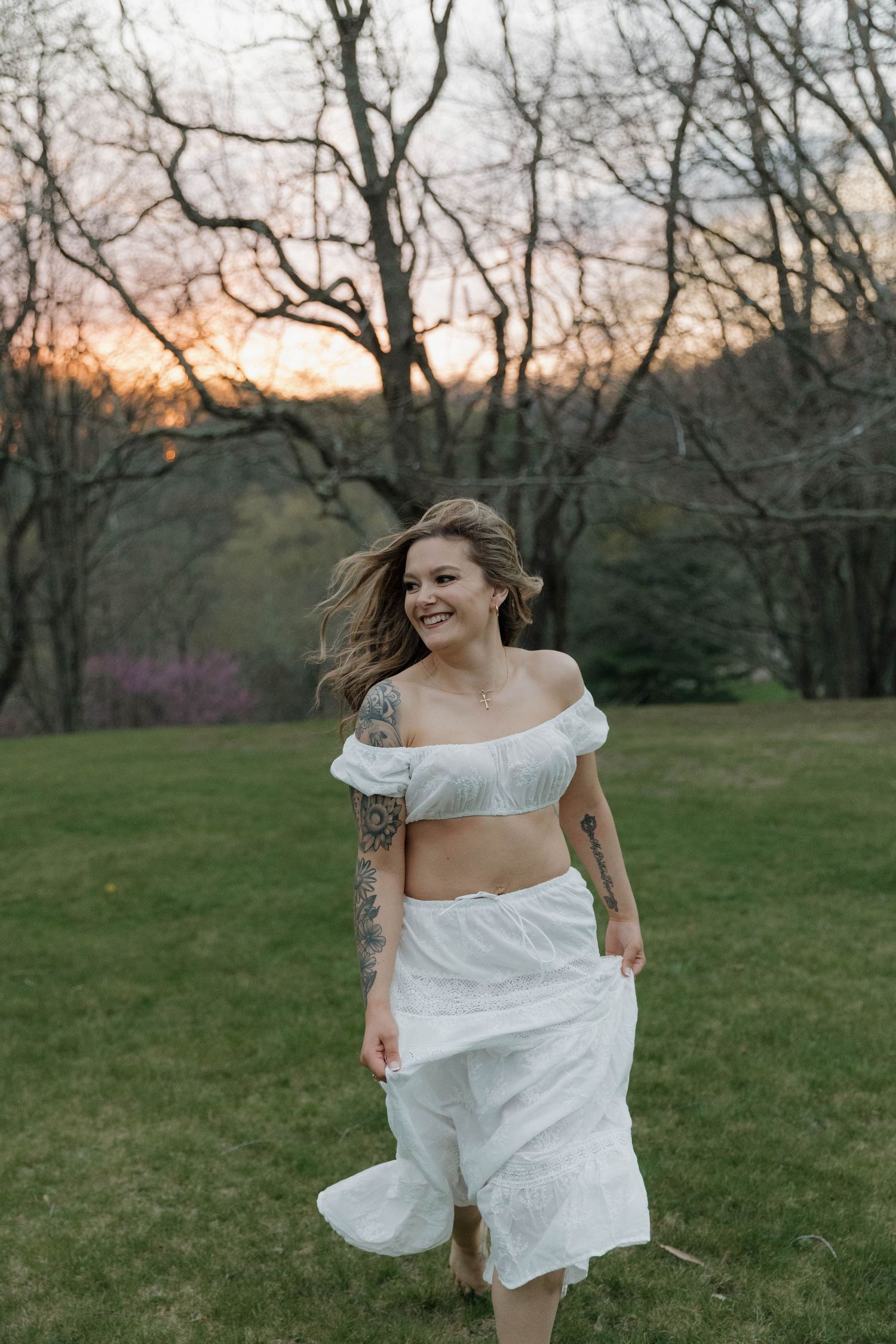 A joyful figure in a flowing white two-piece outfit dances in a grassy field at dusk with bare trees in the background.