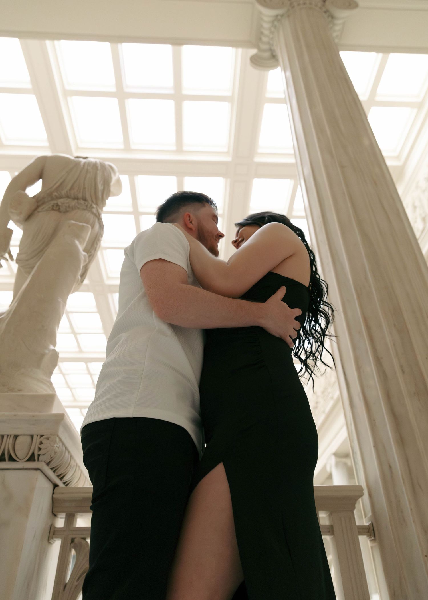 A romantic embrace between two people in formal wear under white draped columns with dramatic lighting.