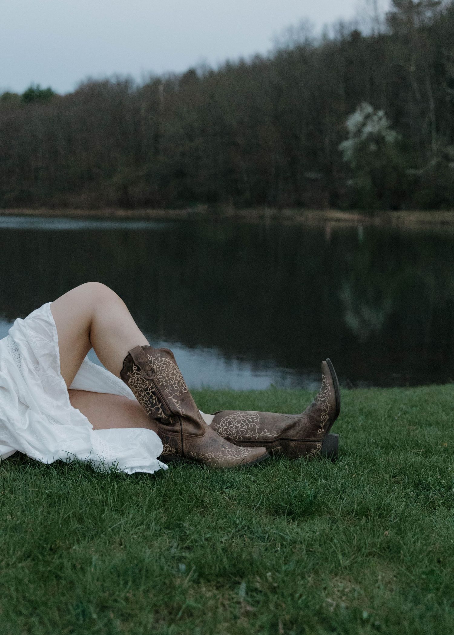 Brown leather cowboy boots with intricate studded details peek out from under a flowing white dress by a serene lake.