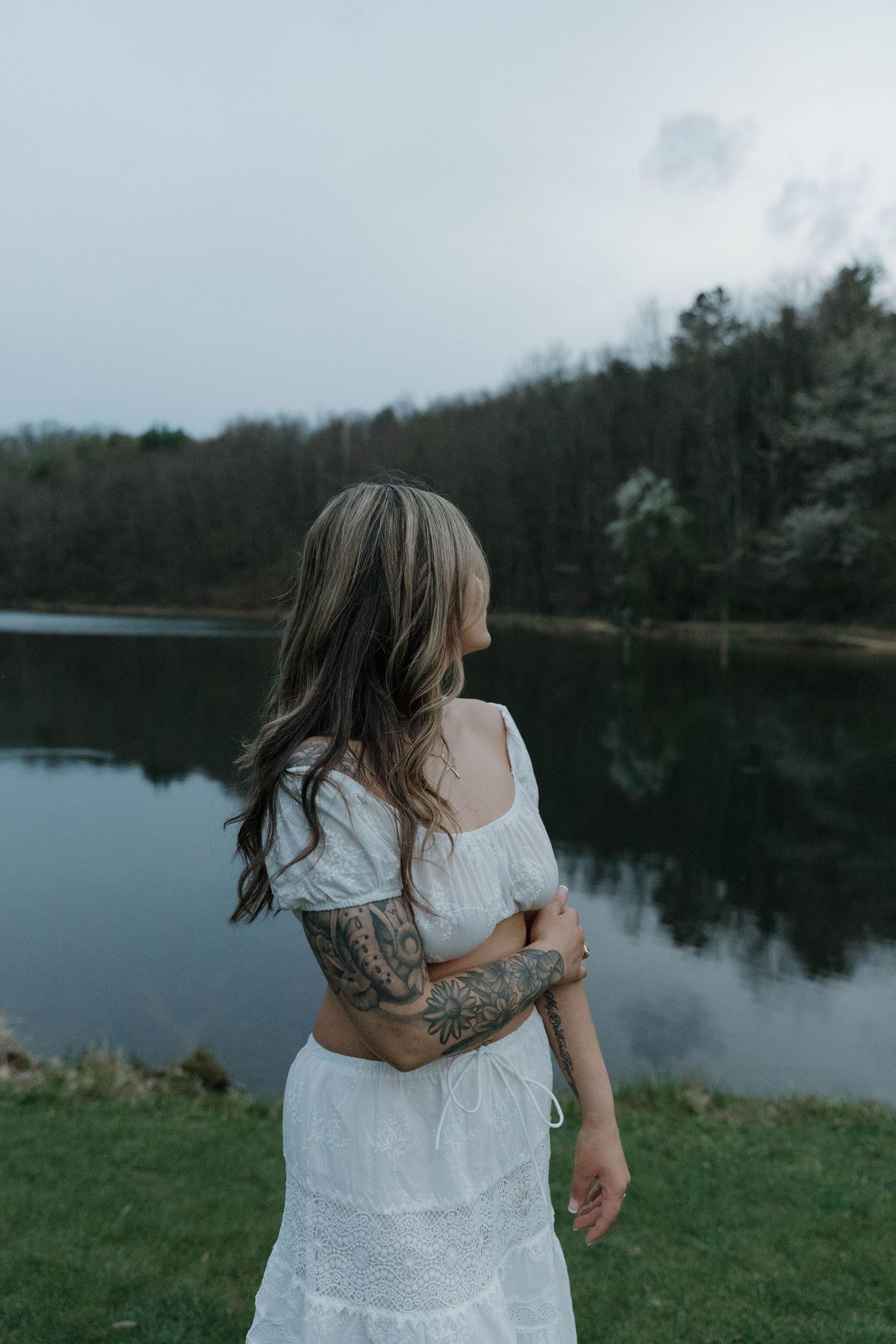 A person in a white dress with tattoos stands by a serene lake at dusk while looking out at the calm water.
