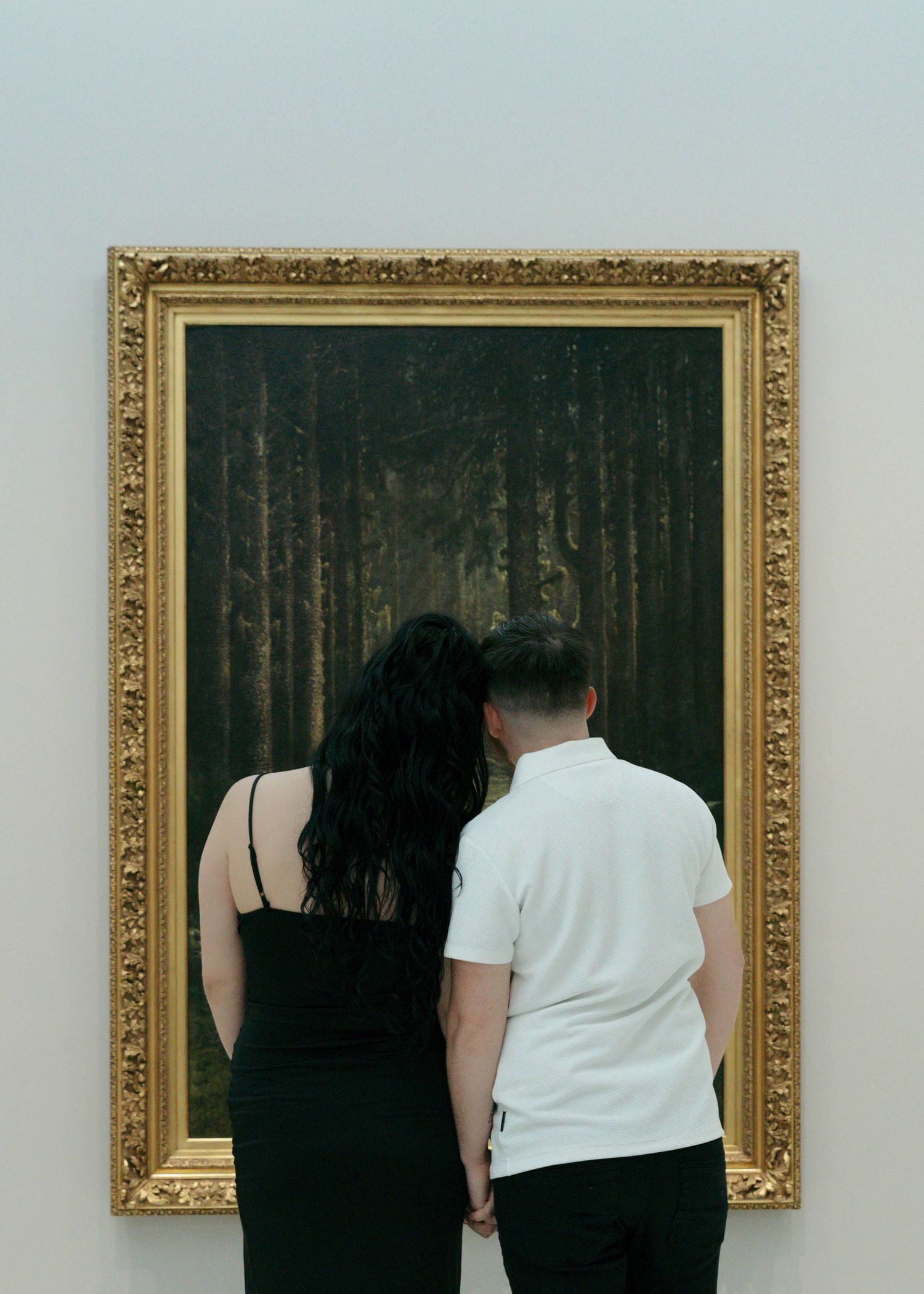 Two people viewing a framed artwork of a dark forest scene in an ornate gold frame at an art gallery.