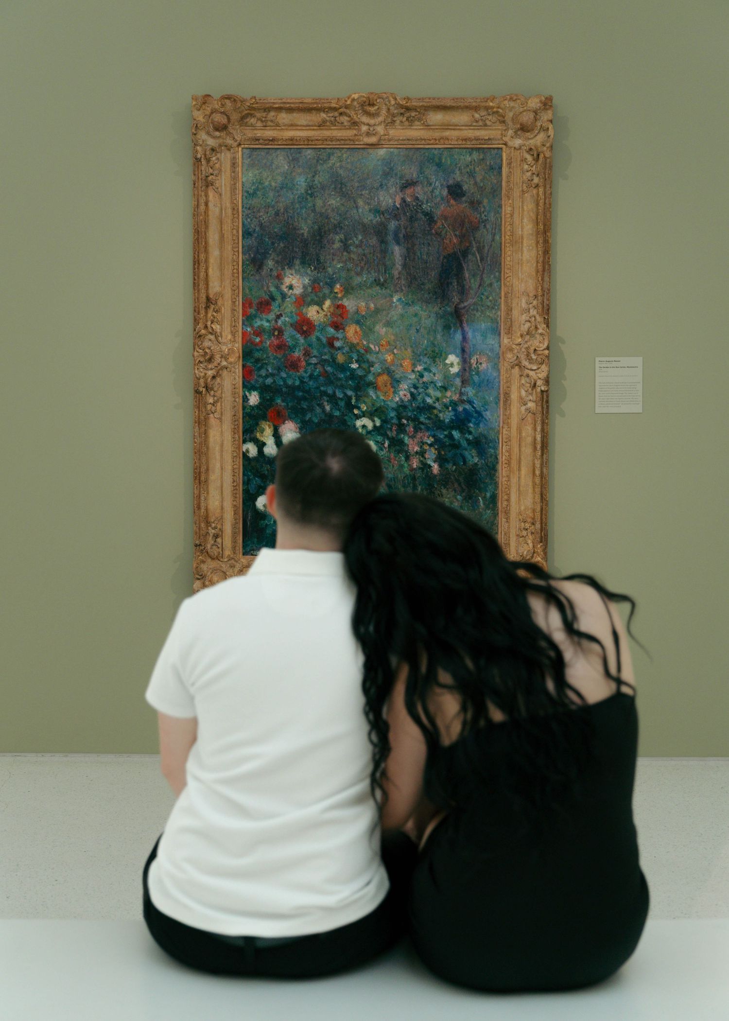 A couple sits together on a bench viewing artwork in an art gallery with ornate gold frames on green walls.