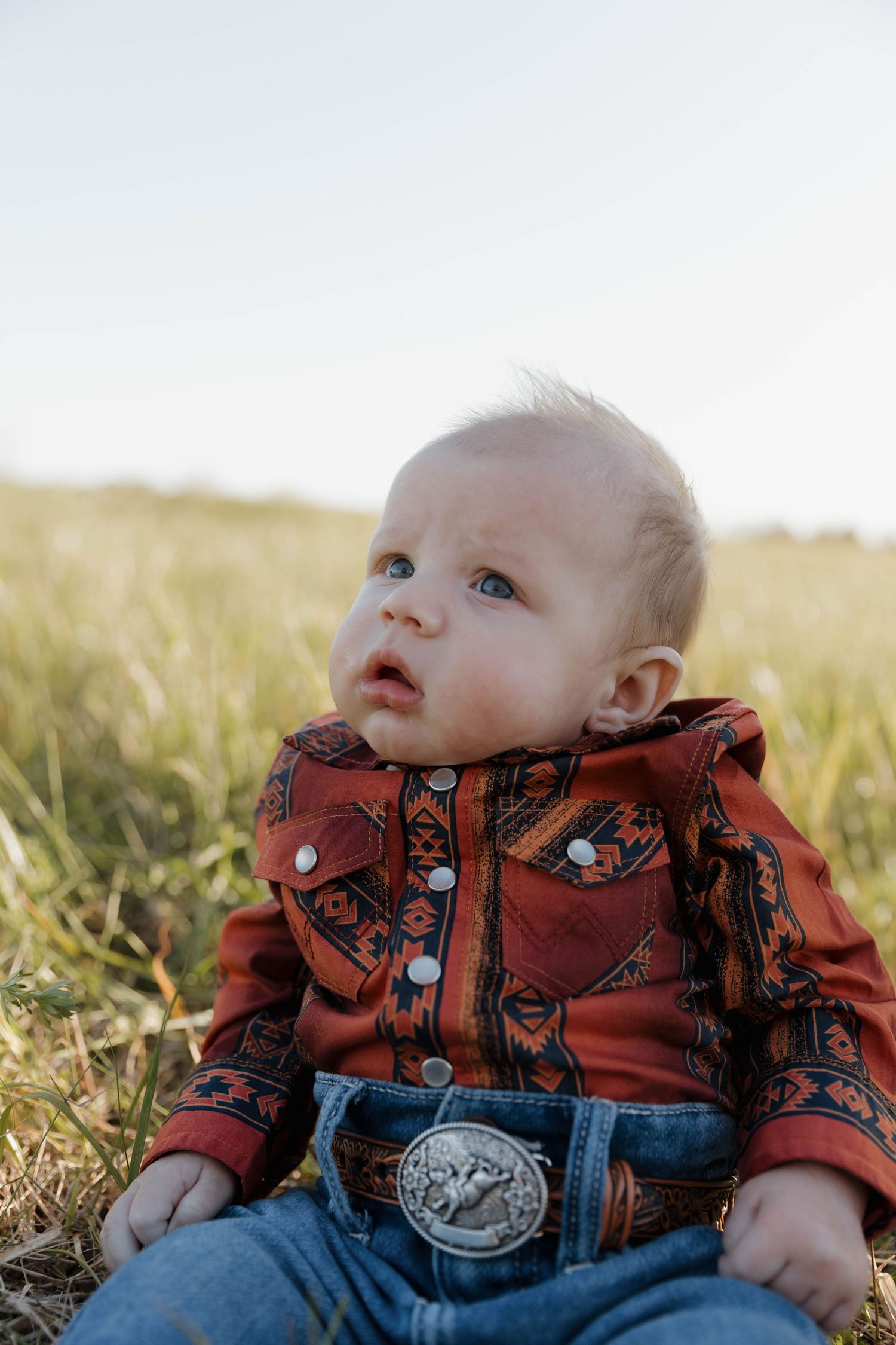 Small baby sitting in grassy field wearing red western shirt with bandana pattern and large belt buckle.