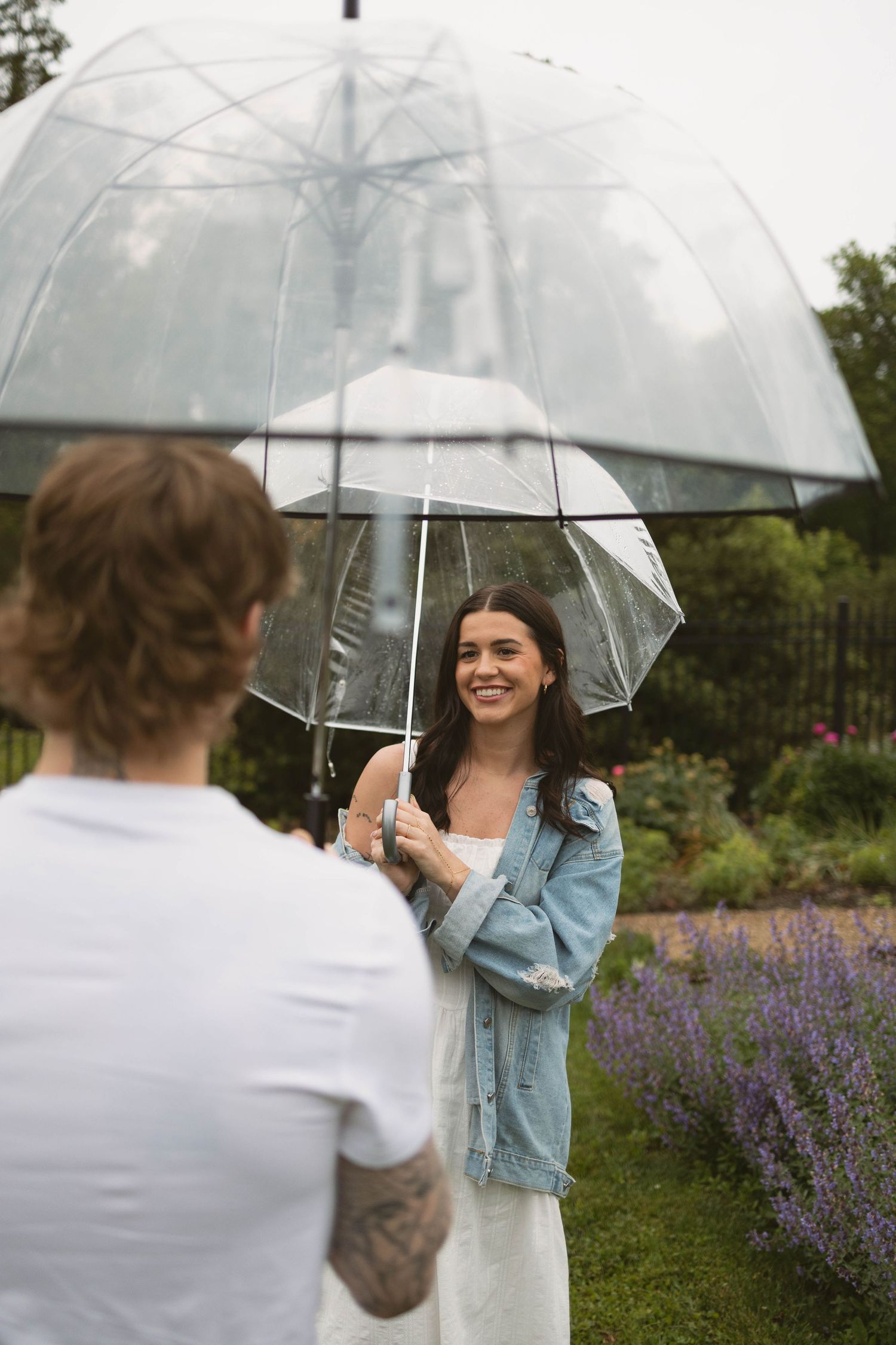 Photographer and model with clear umbrella doing outdoor photoshoot in garden with purple flowers.