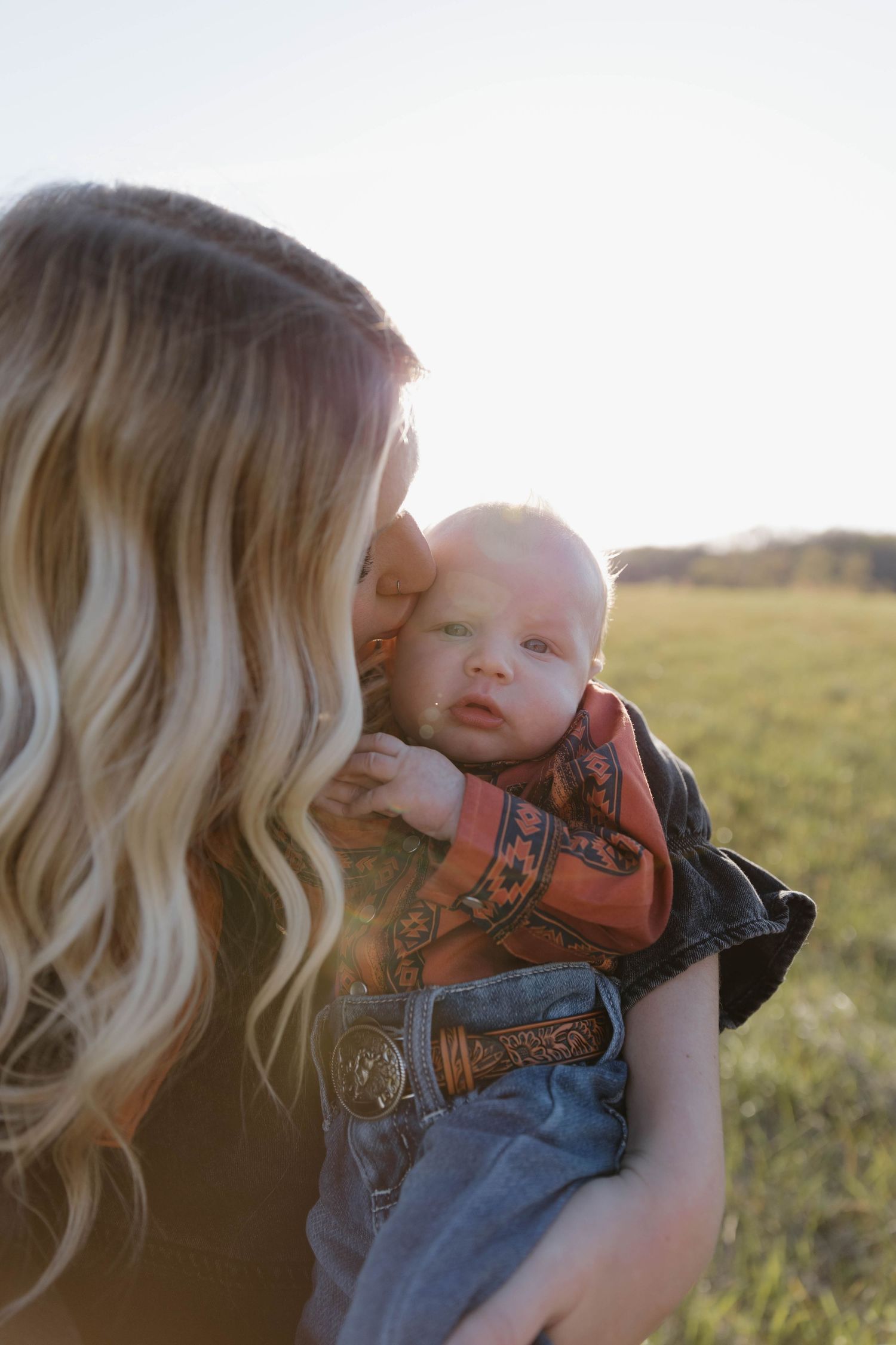 Mother and baby share a tender moment outdoors in a field at sunset, with soft golden light illuminating the scene.