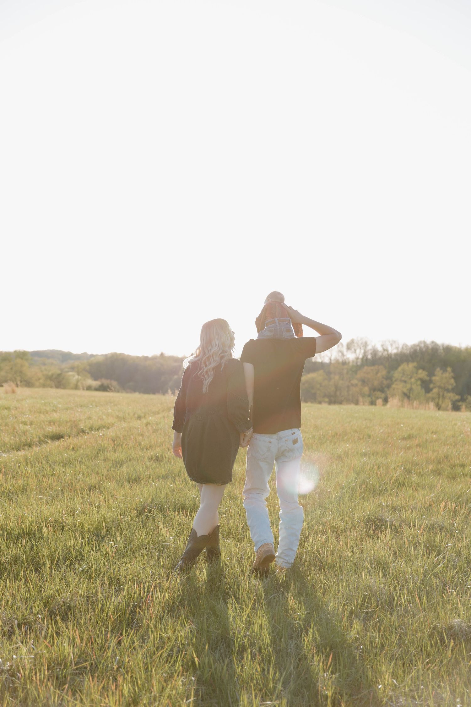 A couple walks together through a sunlit meadow at golden hour with long grass and trees in the distance.