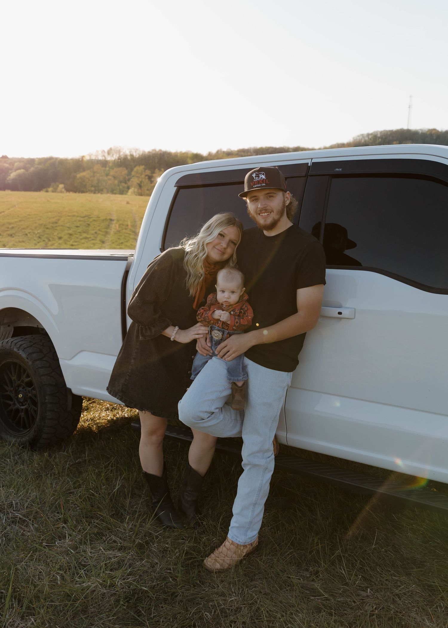 A white pickup truck parked in a grassy field at sunset with a family posing beside the passenger door.
