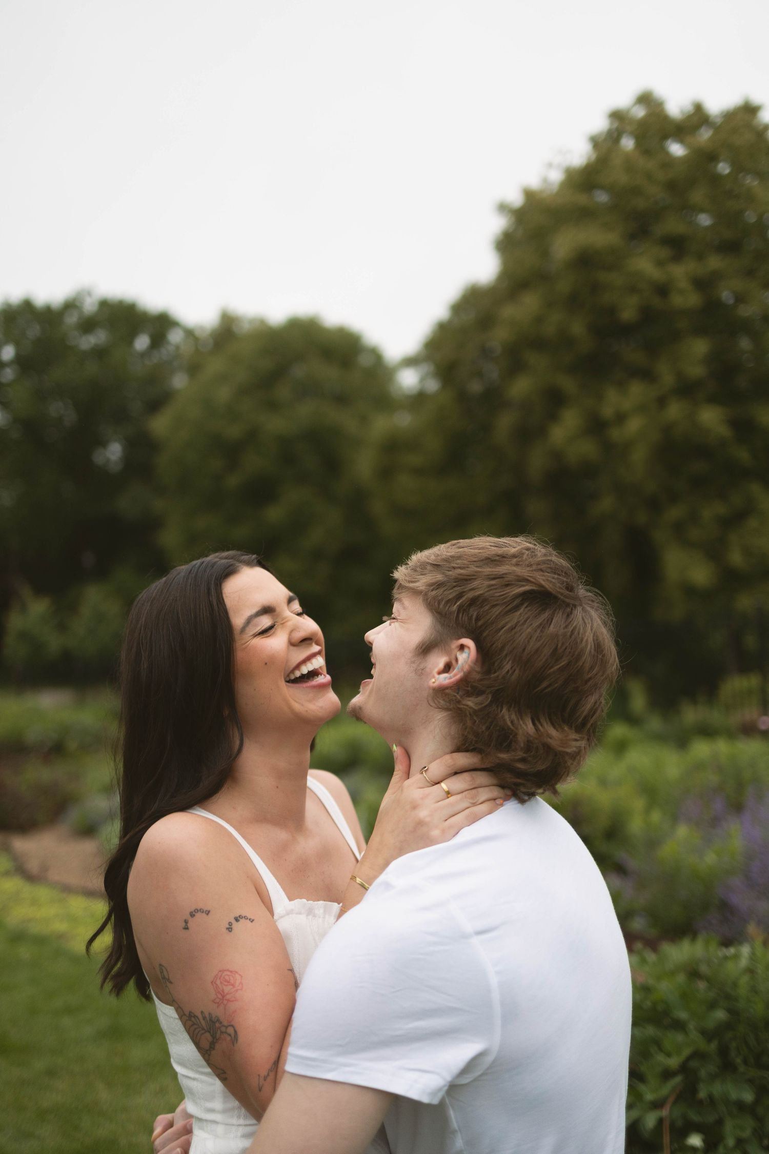 A couple laughs joyfully together in a garden setting with greenery and trees in the background.