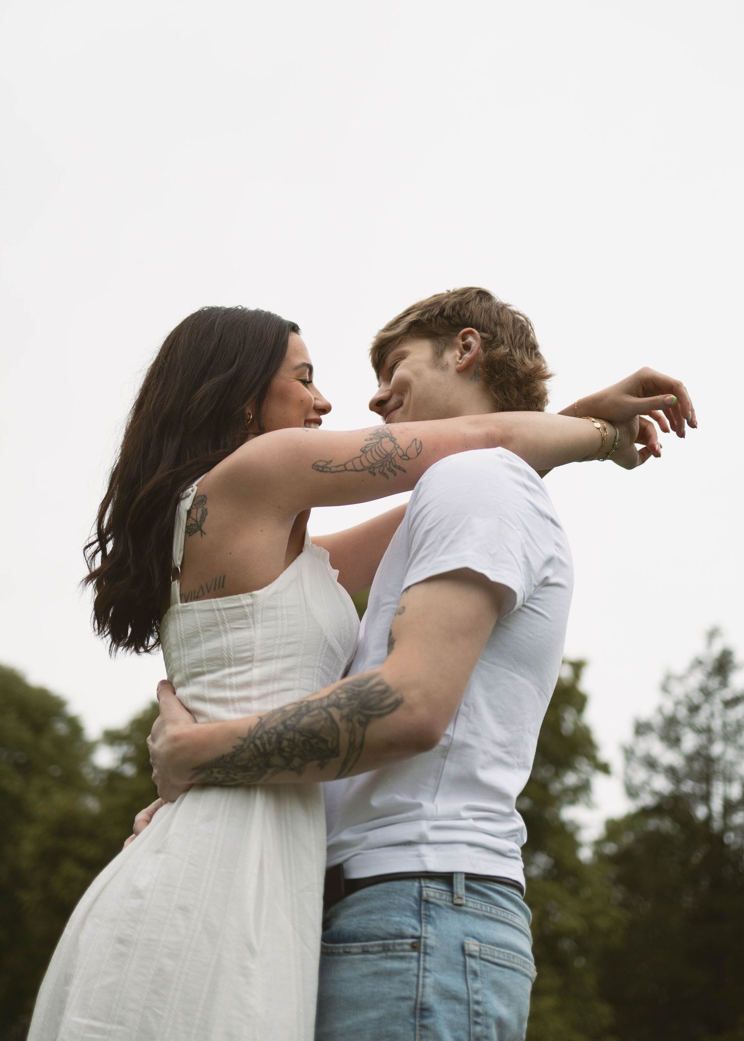 A romantic couple embraces outdoors in summer, with the woman in a white dress and the man wearing a casual t-shirt.