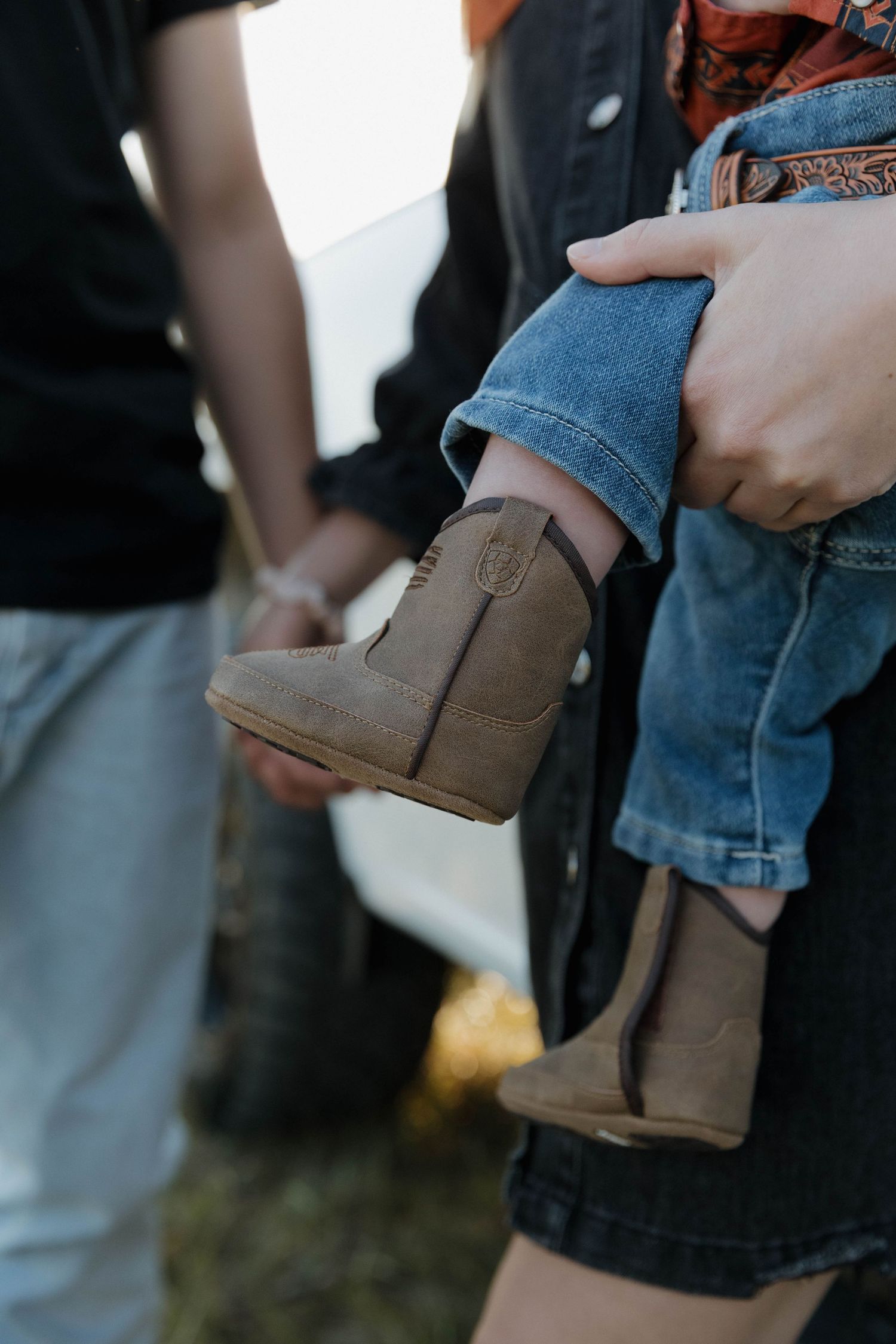 Close-up of small brown leather cowboy boots on child's feet with jeans being held by adult hands.