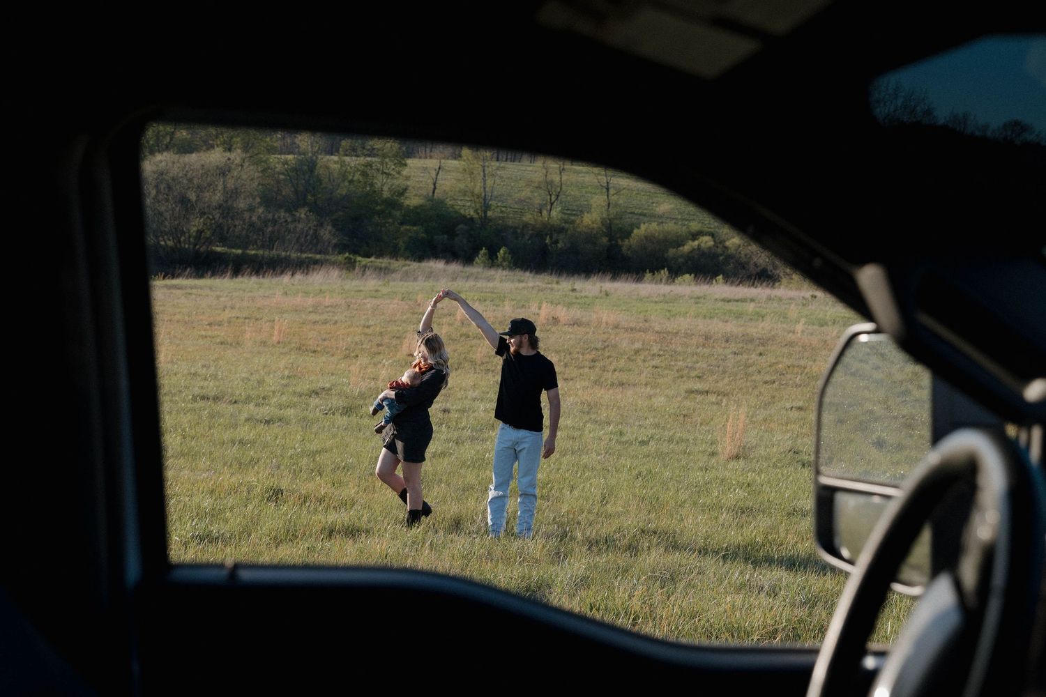 Two figures in a grassy field seen through a car window as they walk together in afternoon light.