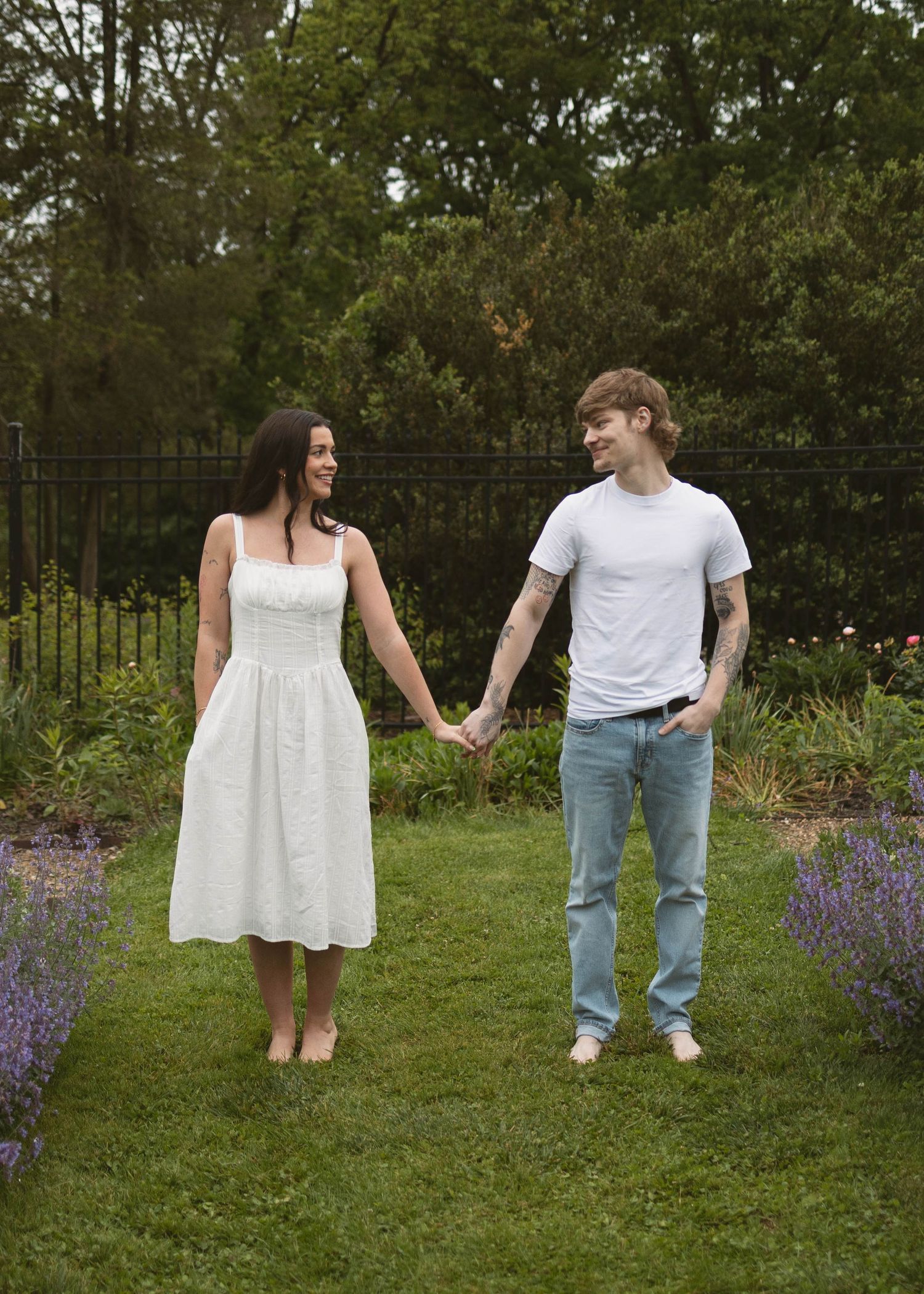 A couple holding hands while standing barefoot in a garden with blooming lavender and greenery.