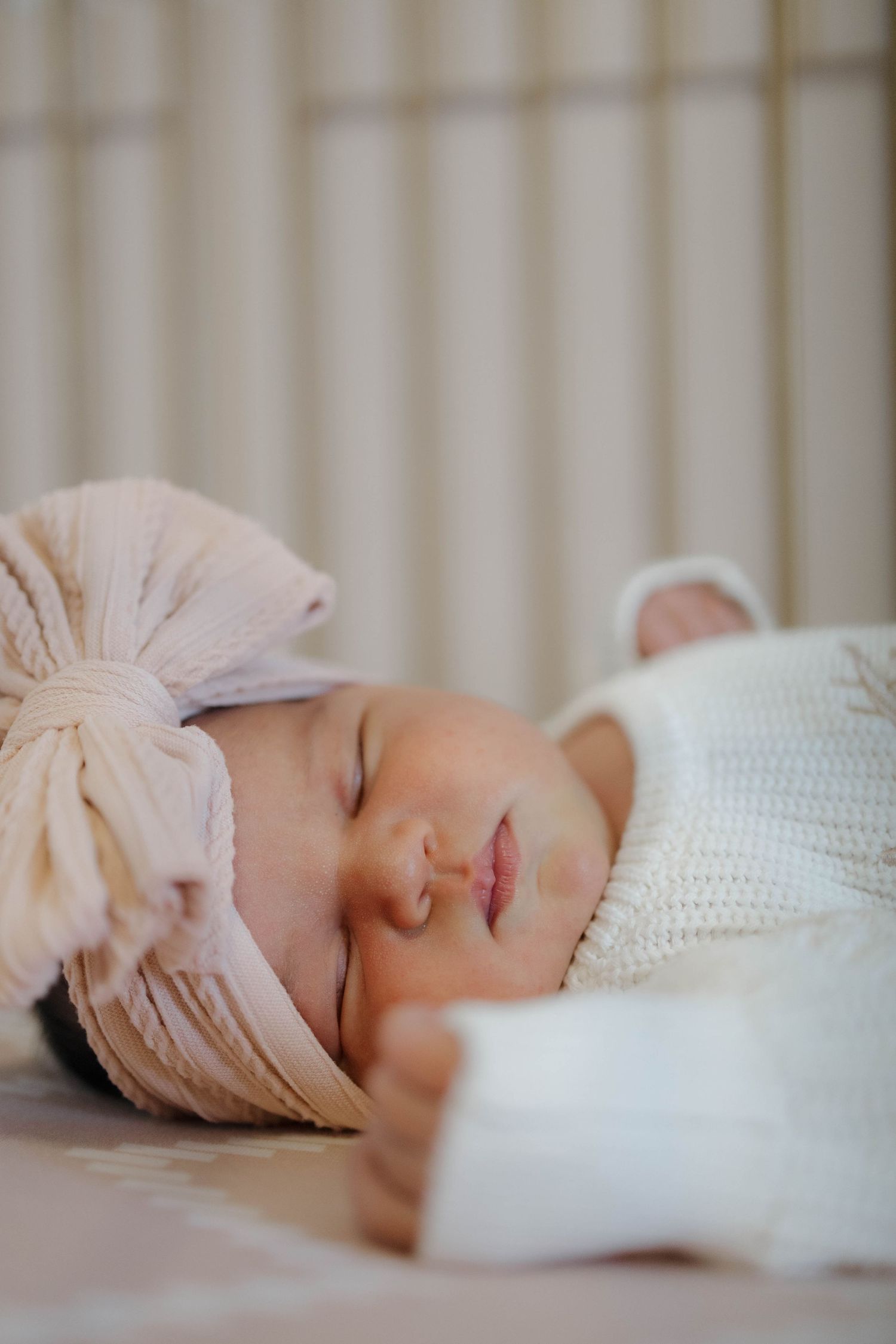 Peaceful sleeping baby wearing pink bow headband and white sweater rests on soft bedding.