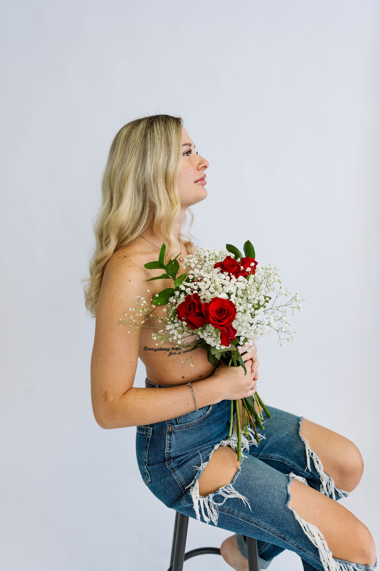 Side view of someone holding a bouquet of red roses and baby's breath while sitting on stool wearing distressed jeans.
