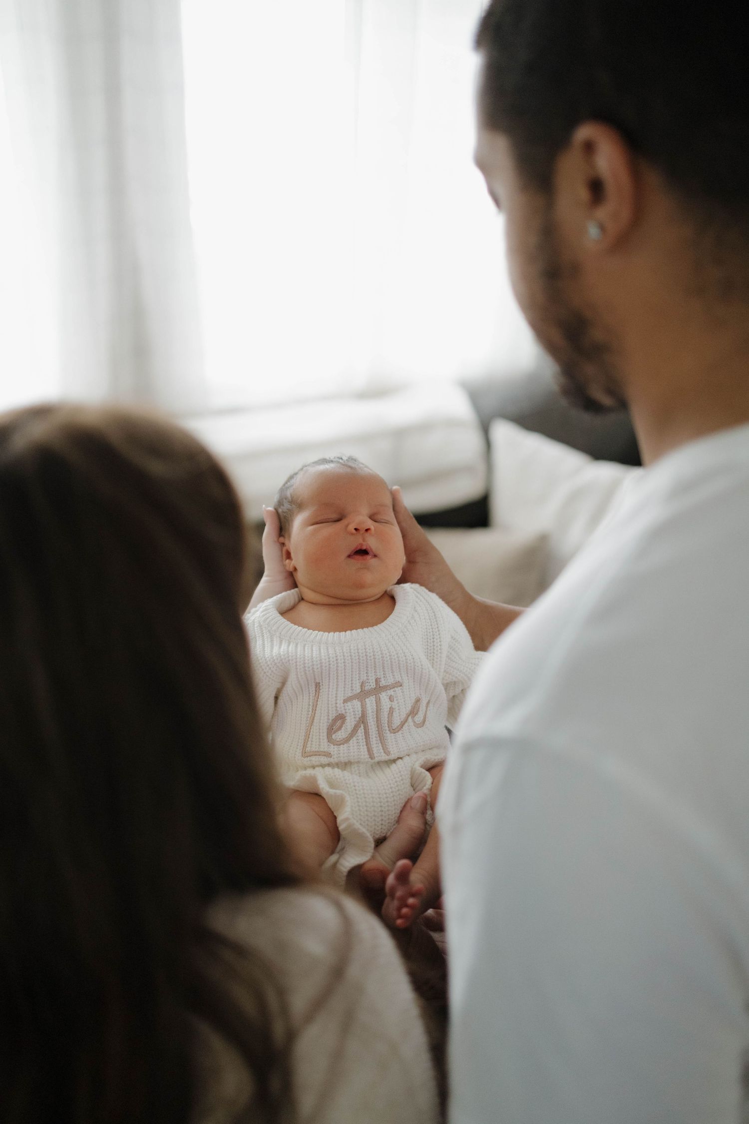 A sleeping newborn baby in a white onesie that says 'little' being held by their parents in a soft sunlit room.