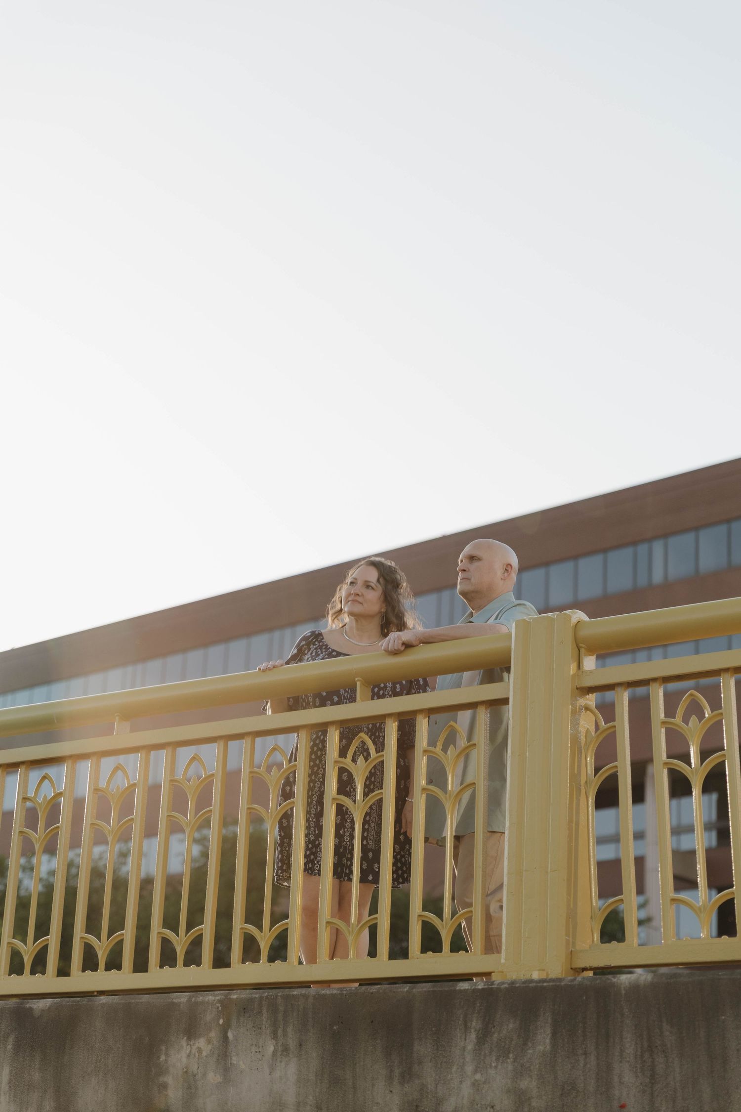 Two people stand together at a golden railing overlooking a cityscape at sunset.