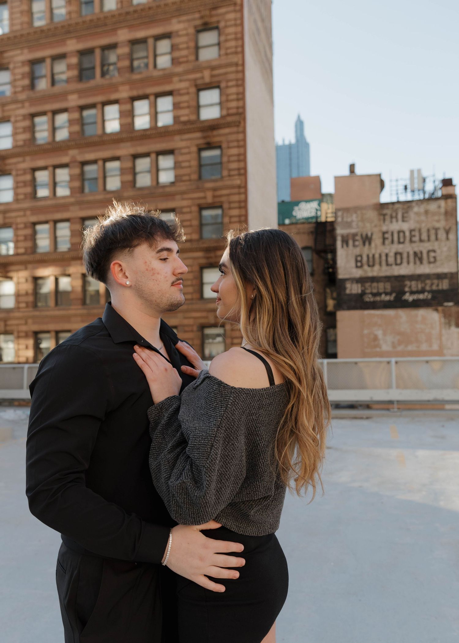 A couple embraces intimately on a rooftop with urban architecture and the New Fidelity building visible in the background.