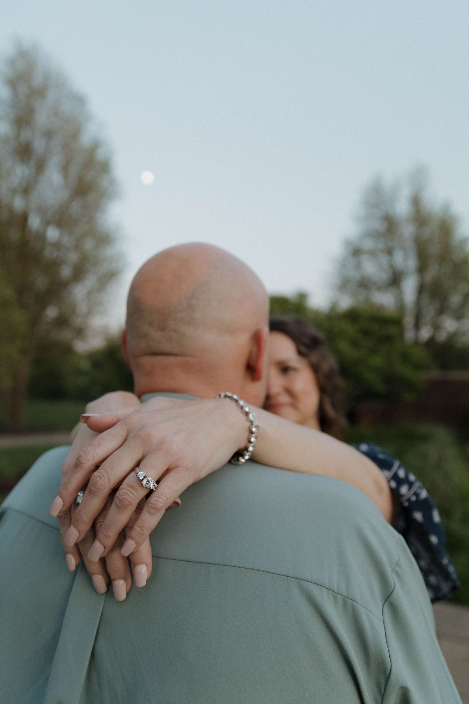 Close-up view of hands with engagement ring embracing during intimate outdoor sunset moment with moon visible in sky.