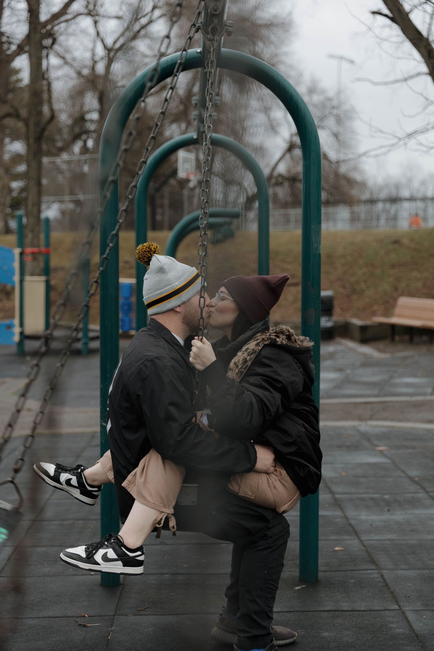 Two people share a tender moment on a playground swing on a cloudy winter day, both wearing warm clothes and sneakers.