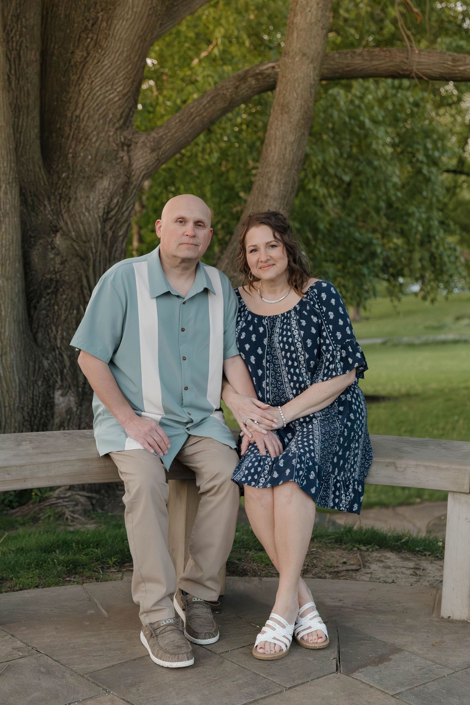 A couple sits together on a stone bench in a garden setting wearing casual summer attire.
