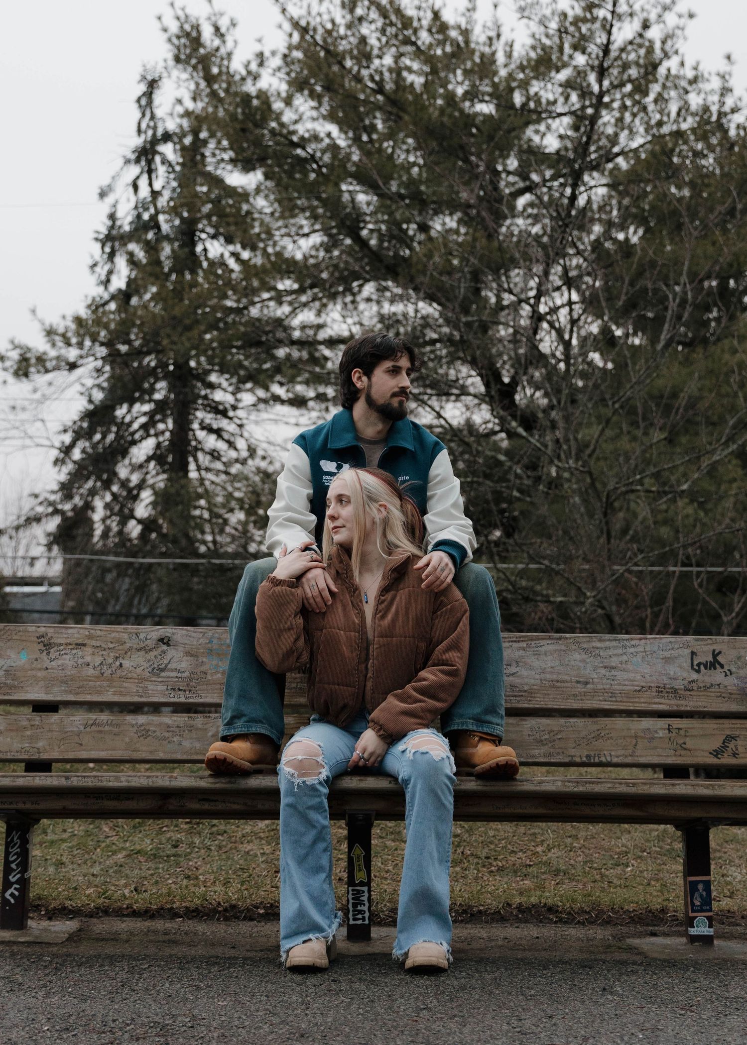 Two people sitting together on a park bench surrounded by trees on an overcast day.